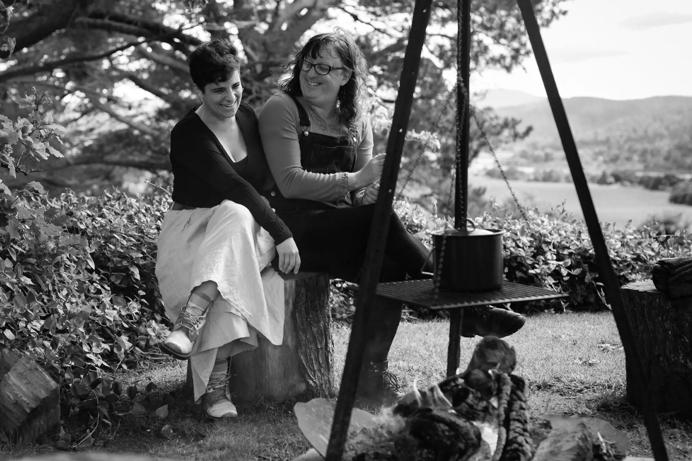 Two women sit closely on tree stumps in a garden, under a large leafy tree. One wears a cardigan and skirt, the other overalls. They are smiling and appear to be sharing a joyful moment, with a campfire tripod holding a pot visible in the foreground