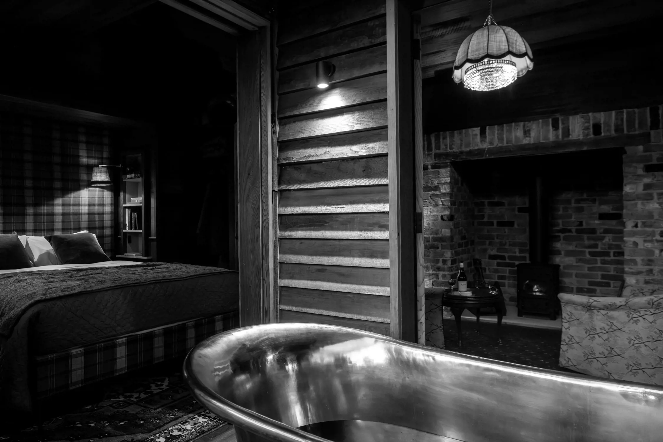 A warm and cozy rustic interior. A metal bathtub gleams in the foreground, while a plaid-covered bed with reading lamps sits in a nook. A brick-walled room with a wood-burning stove and soft lighting completes the scene, suggesting relaxation and comfort.