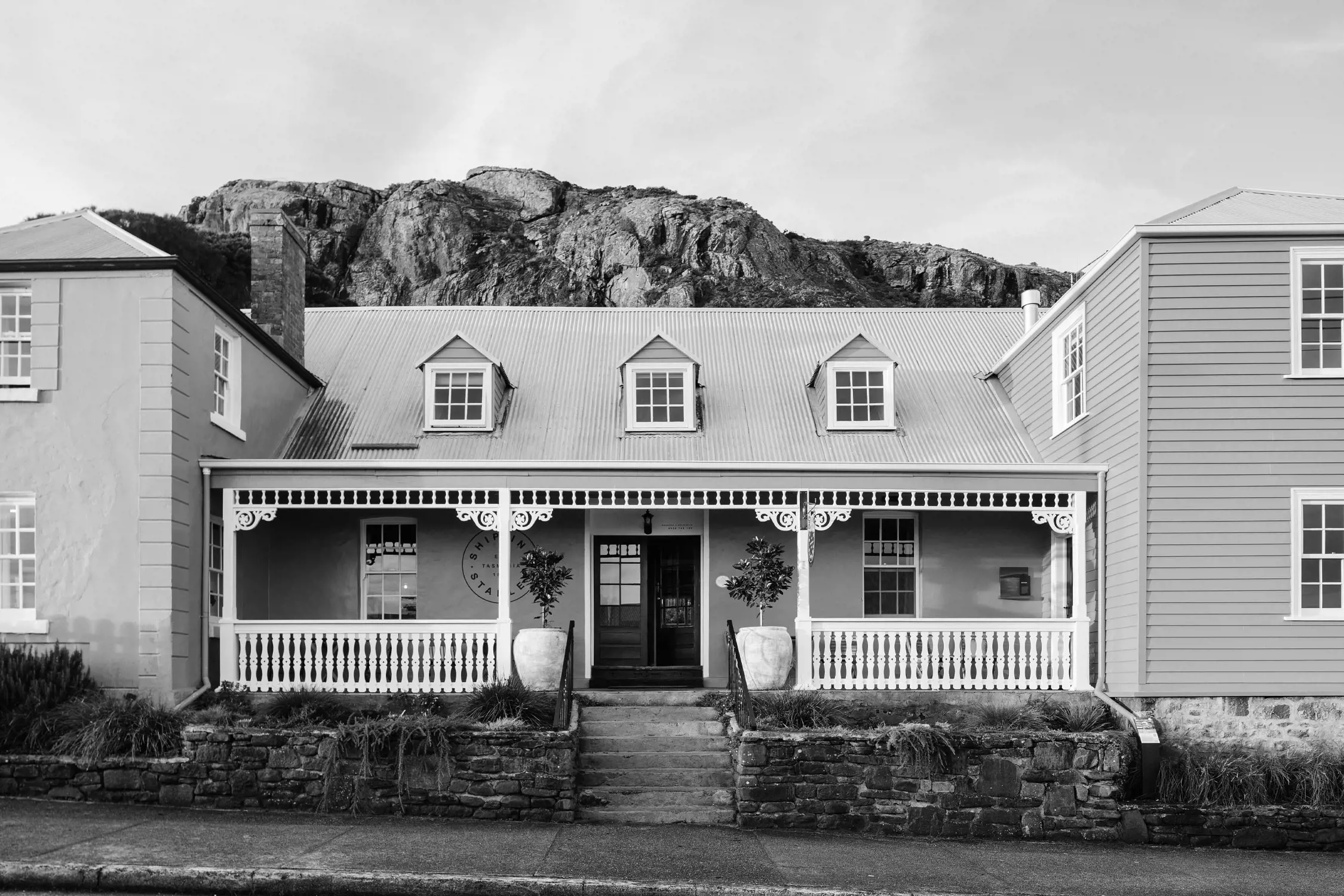 A charming colonial-style inn with a symmetrical facade painted in soft pastel tones. The building features a gabled roof, a veranda with intricate wooden trim, and a lush garden in front. The dramatic backdrop of a rocky cliff completes the picturesque scene.