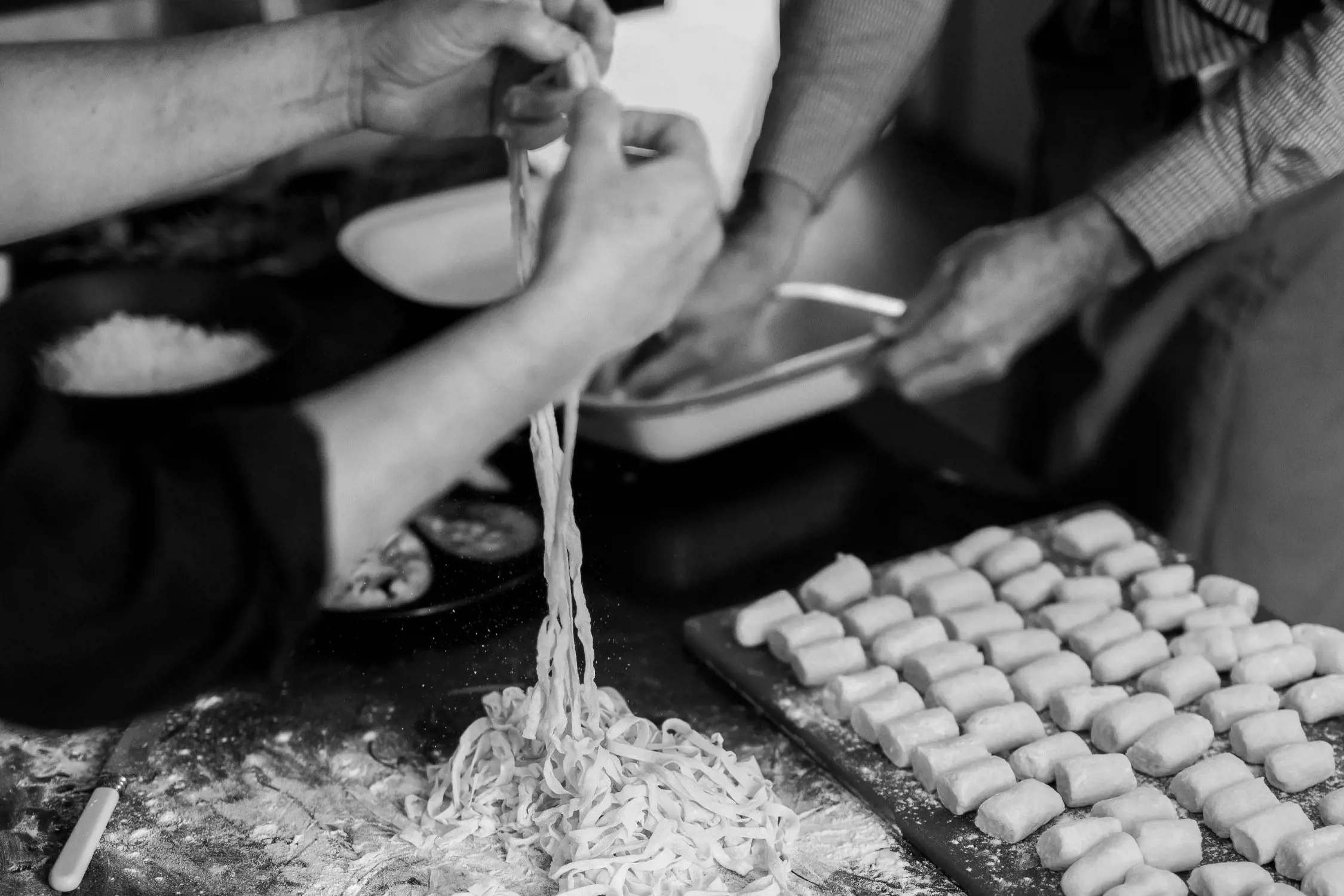 A close-up of hands working in a rustic kitchen. One person stretches fresh handmade pasta, while another shapes small gnocchi pieces, lined up on a floured surface. The table is covered in flour, creating an atmosphere of creativity and culinary craftsmanship.