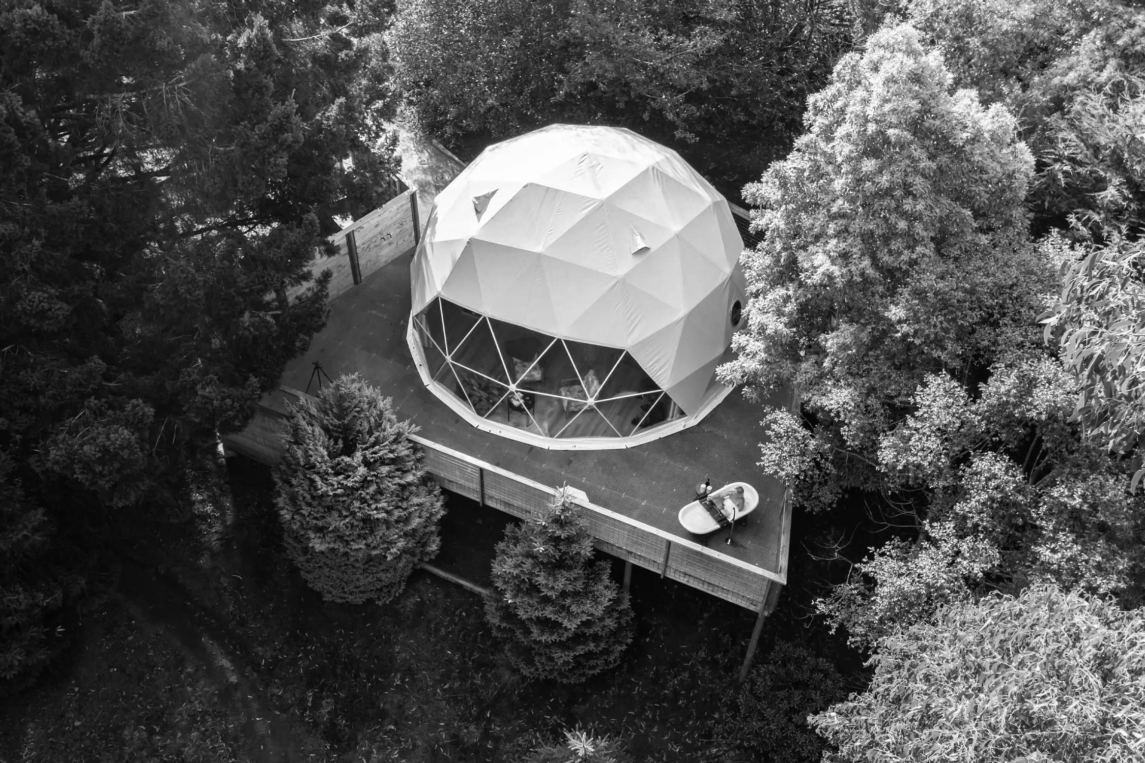 A domed white structure, resembling a geodesic glamping pod, nestled amidst tall trees in a serene forest. A wooden deck surrounds the pod, and a small outdoor bathtub is visible on the side, offering an inviting retreat in nature.