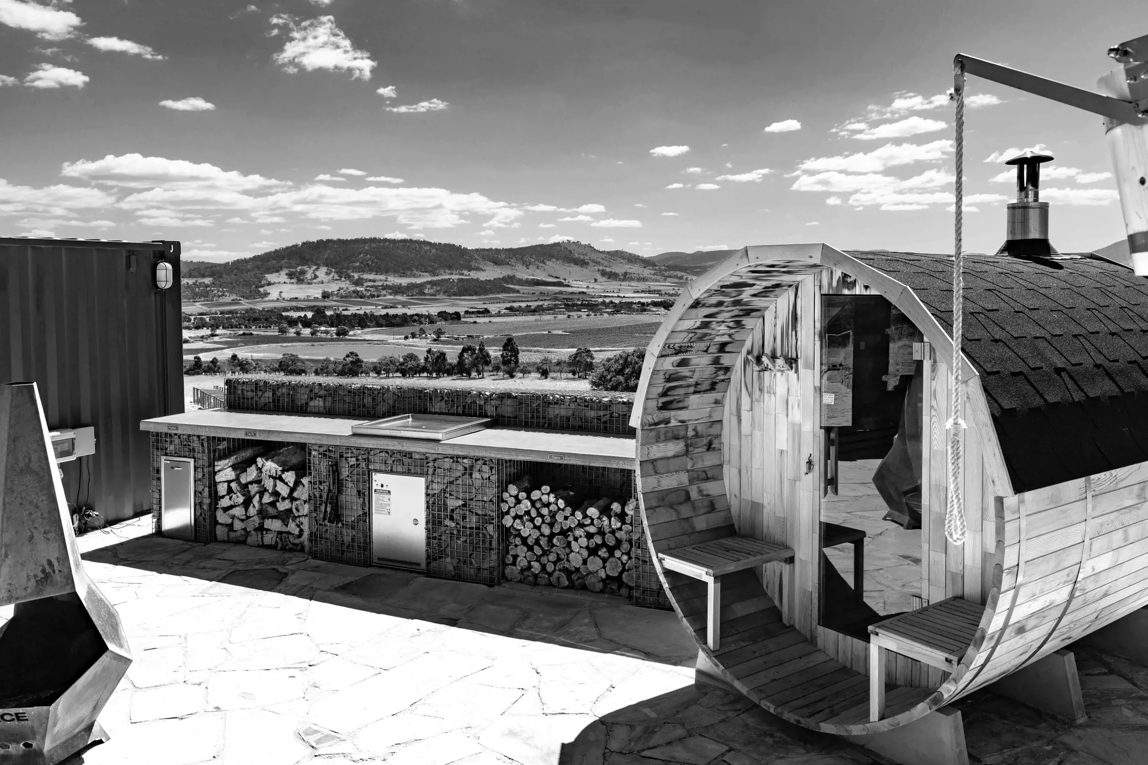 An eco-friendly retreat surrounded by dense greenery. A unique dome sauna with a chimney sits on a sandstone platform. In the background there are rolling hills, green fiels, trees and a dam. 