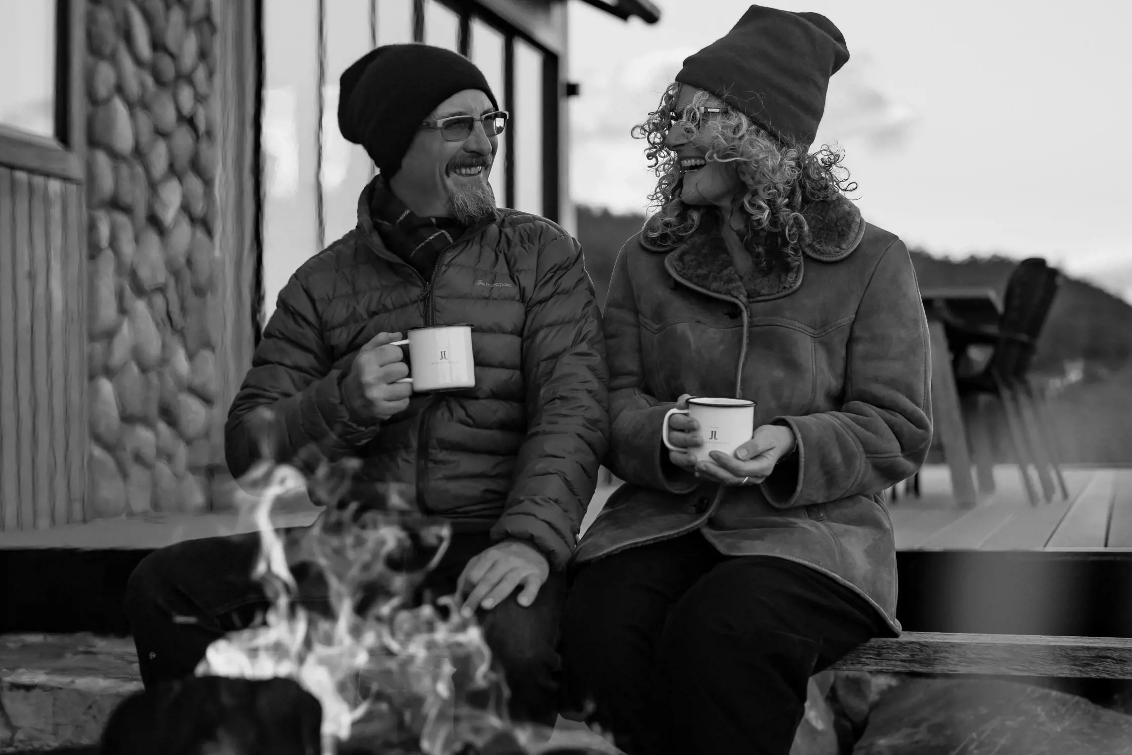 A man and a woman sit together by an outdoor fire pit, enjoying hot drinks from mugs. Both are dressed warmly, with jackets, scarves, and beanies, as they smile at each other in the cool evening light. Behind them, a wooden building with a stone façade is visible, along with a deck area and distant hills under a soft sky.