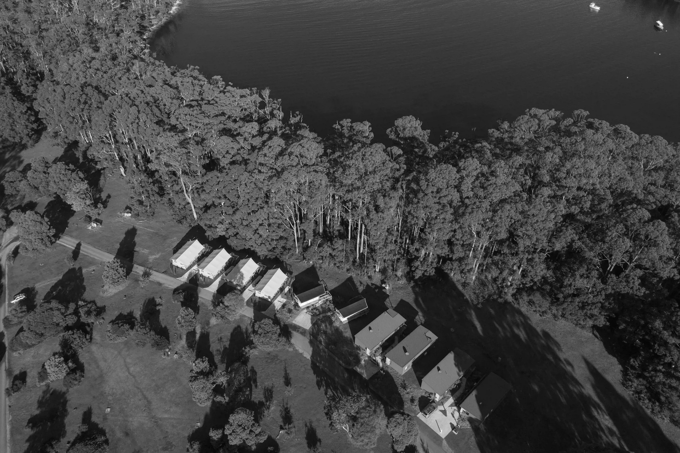 An aerial view of a serene holiday park nestled beside a calm body of water. A row of neatly aligned cabins with light-colored roofs is visible, surrounded by lush greenery and tall trees. The dense forest creates a natural boundary between the park and the water, while a small road weaves through the landscape. Boats can be seen floating on the water, adding to the tranquil setting.