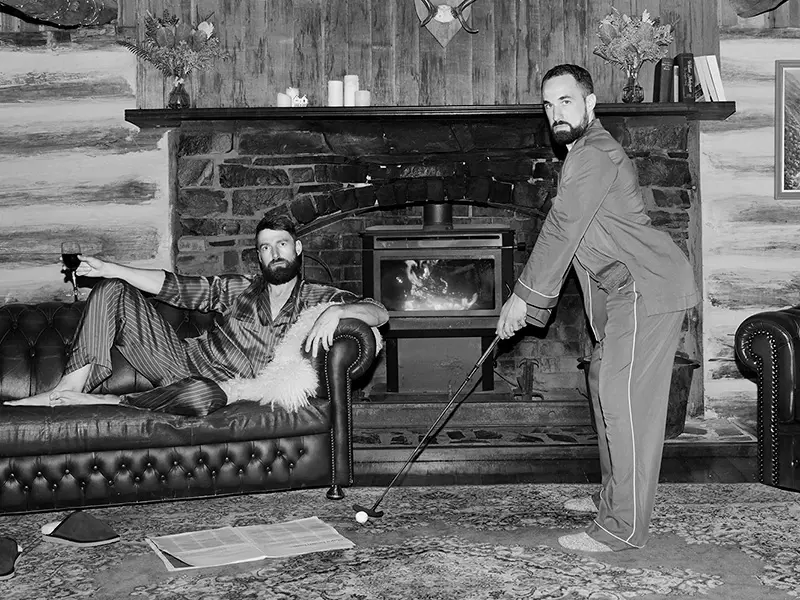 Inside a richly decorated wood log cabin, with a burning fireplace, leather couches and a rich rug. One man lounges on a sofa with a glass of wine while the other plays minigolf - both in bare feet and silk pajamas.