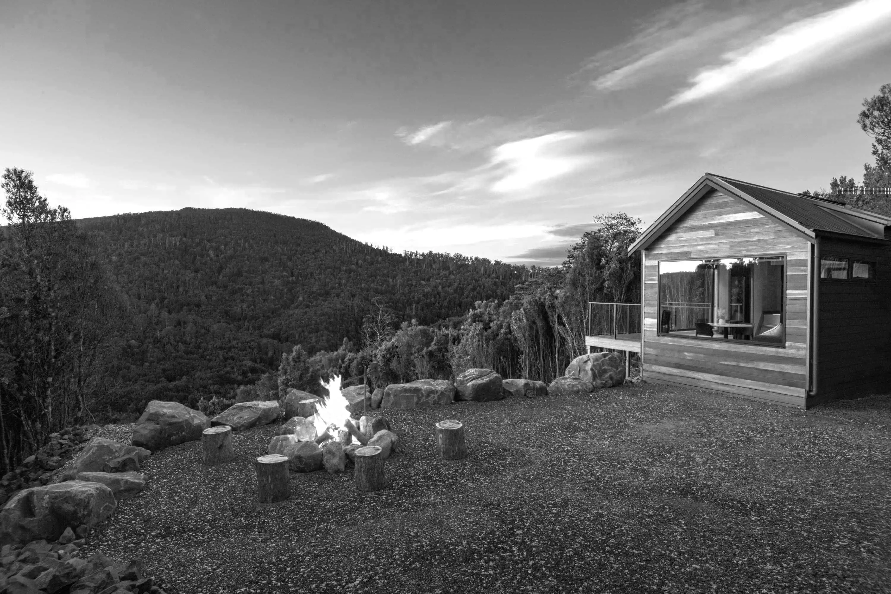 A small wooden building with a fire pit nearby stands on the top of a hill, looking over a forest valley.