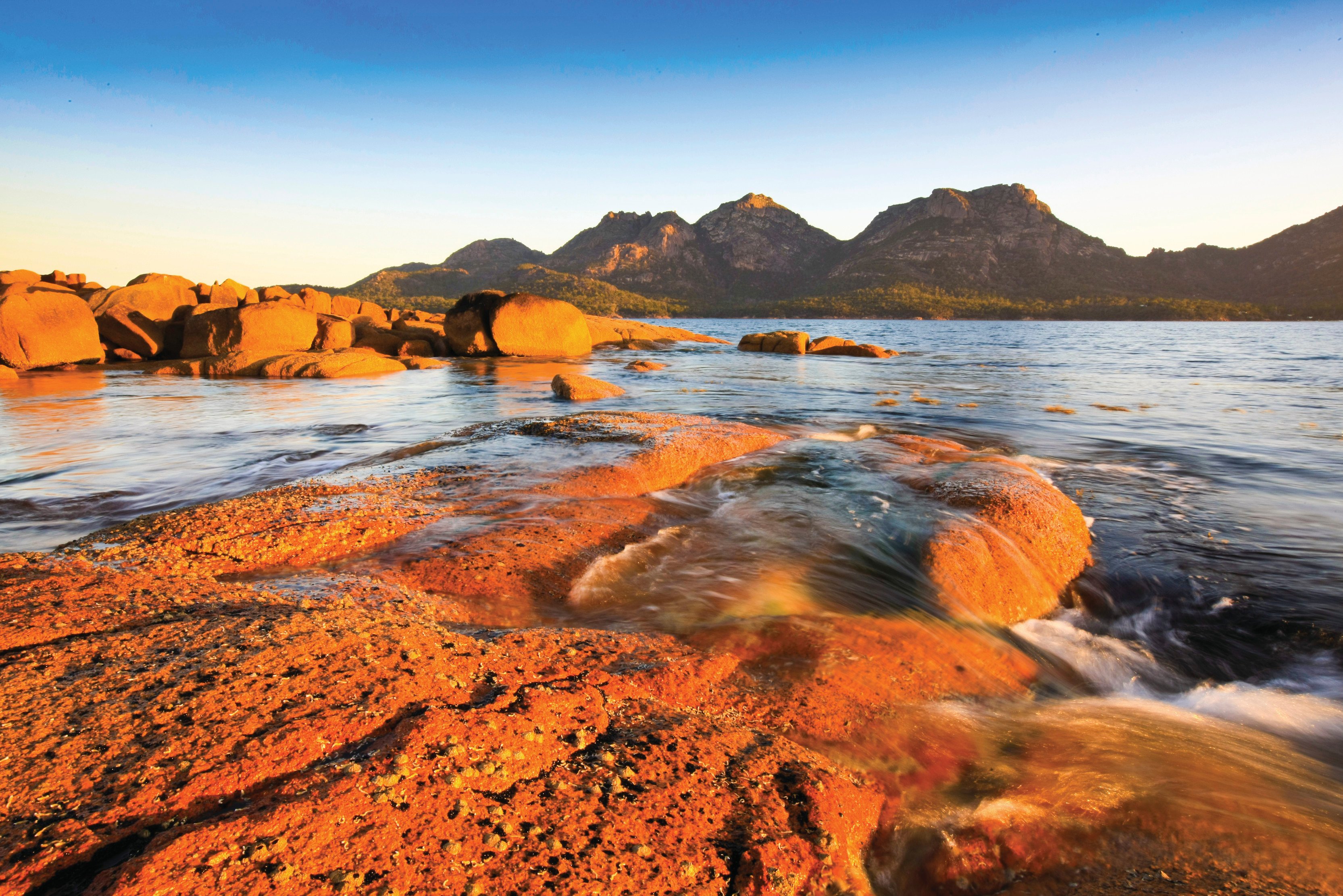 View of the The Hazards in Coles Bay. The rocks shine red as the sun sets over the blue lake.  