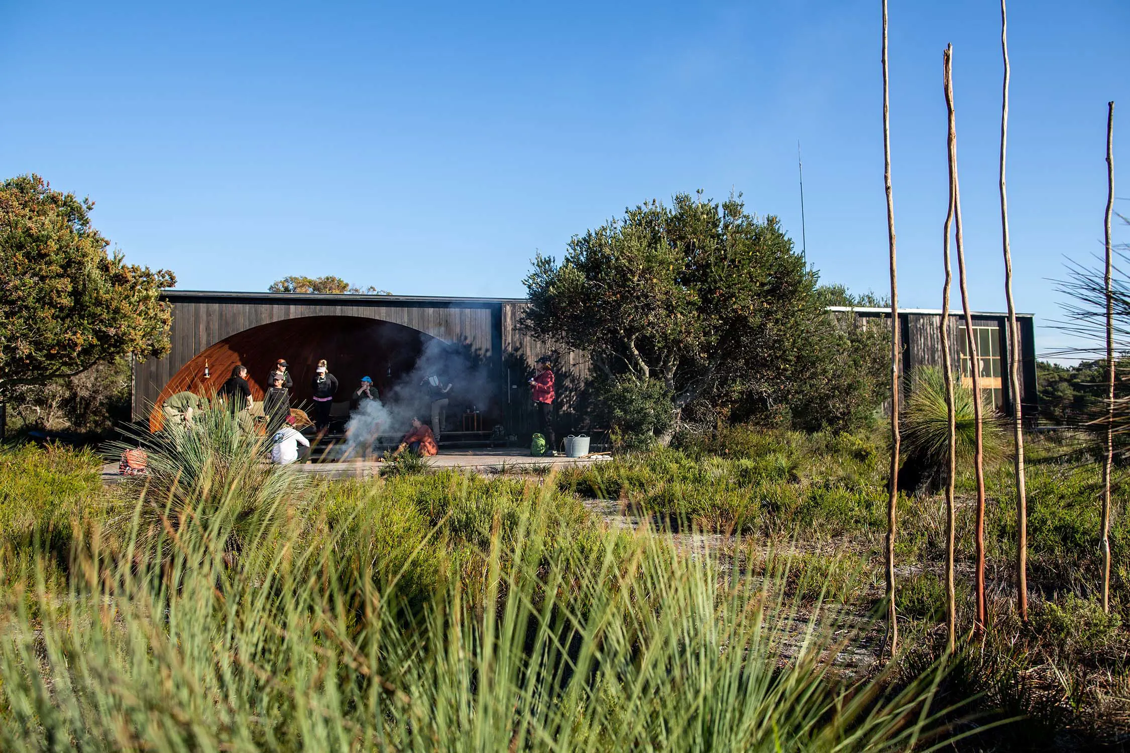 A low-profile wooden building sits amongst shrubs and trees, while a group of people sit in a circle around a campfire nearby.