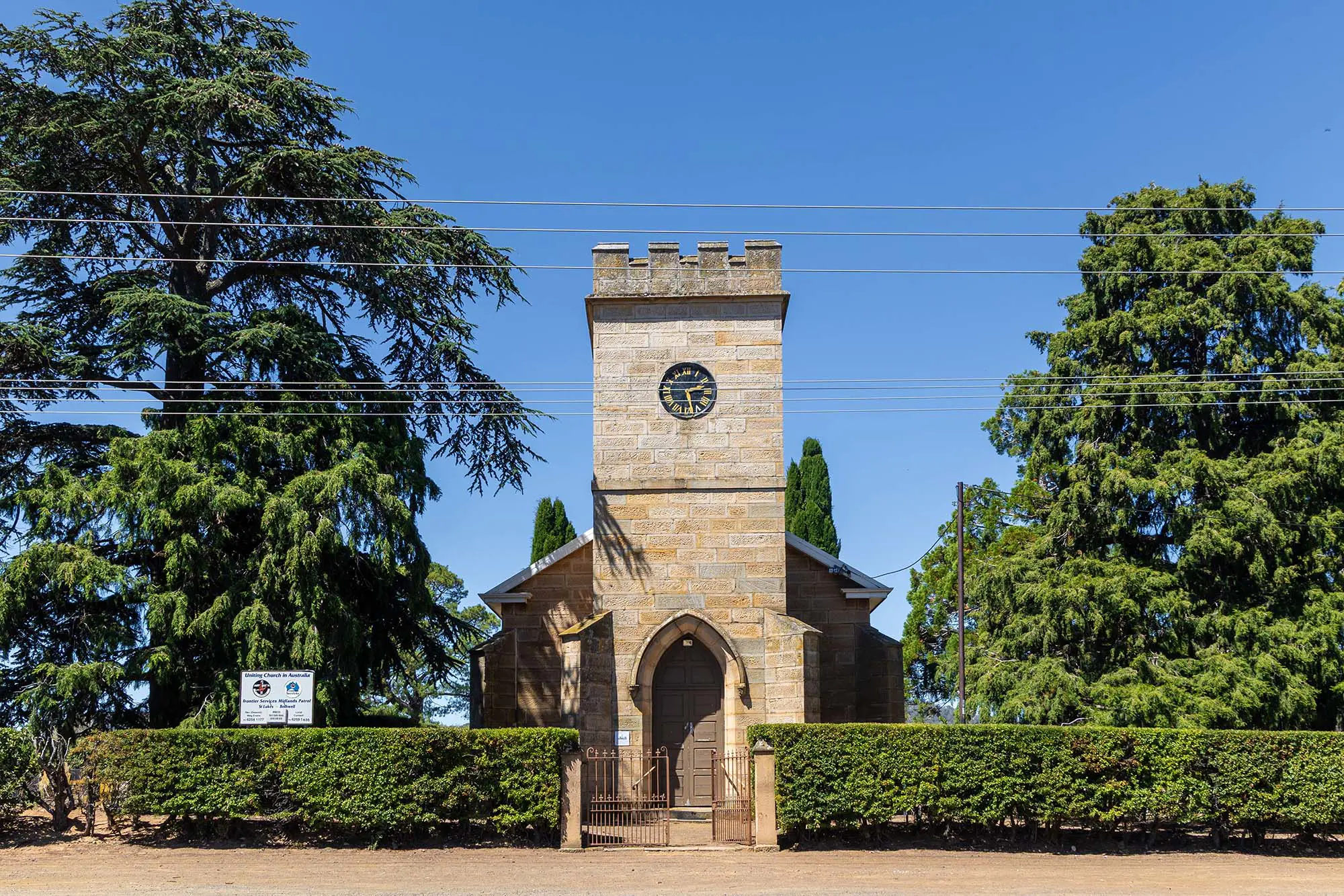 A picturesque stone church tower with open doors and a clock, framed by neat hedges, tall green trees and a clear blue sky.
