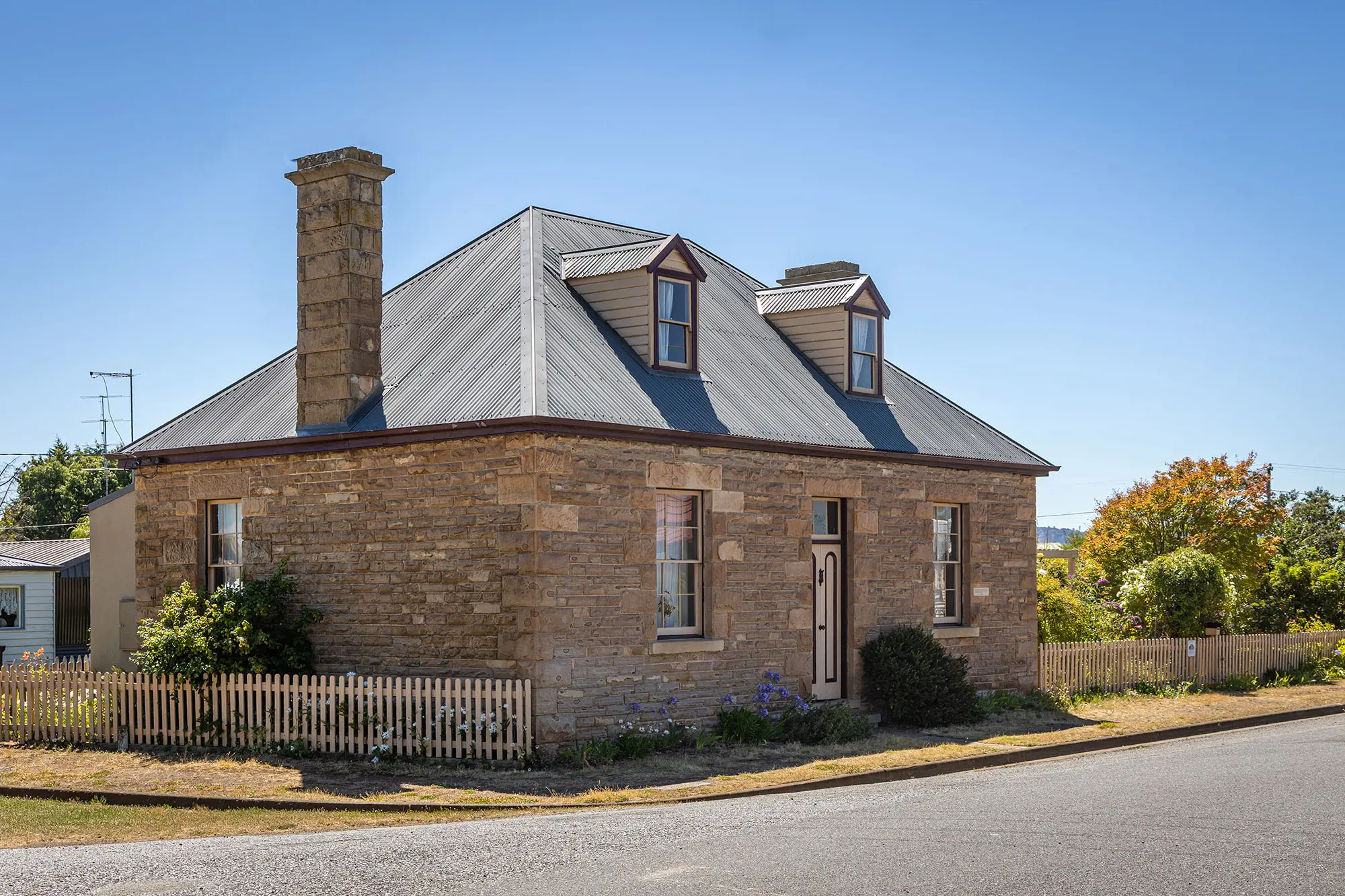 A neat, historic brick cottage, with a dark tin roof and chimney standing tall. The garden and white picket fence are neatly manicured and the sky shines clear blue.