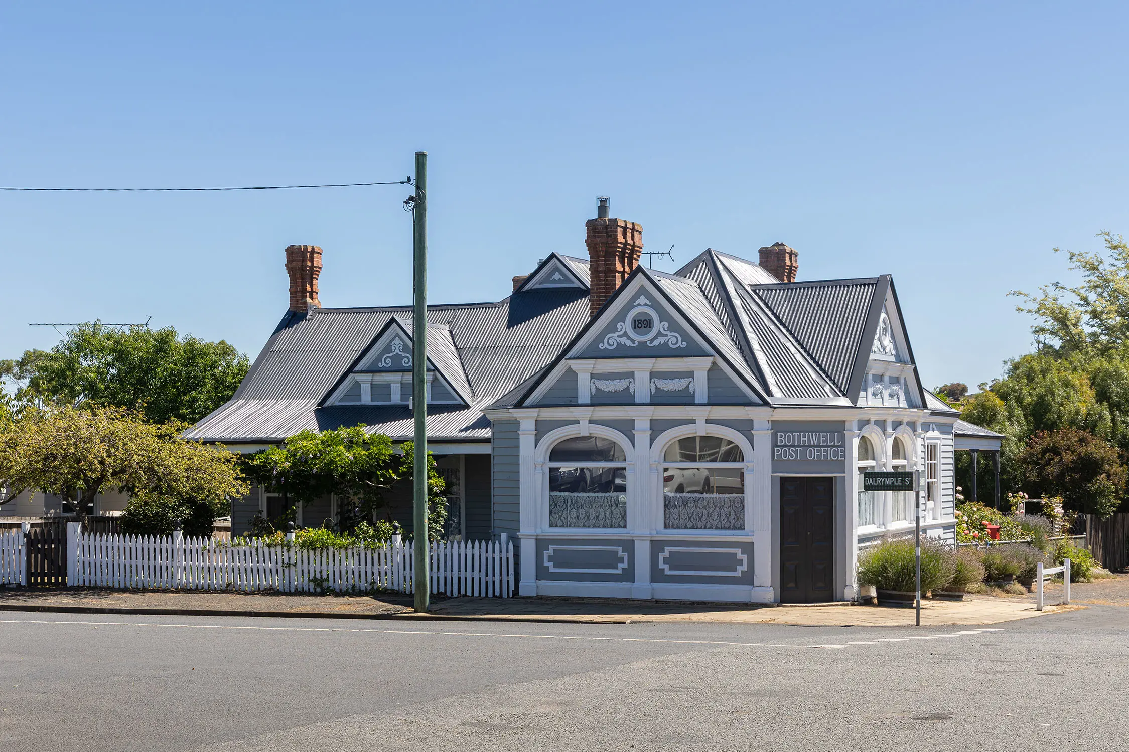 A blue-grey and white historic cottage, with lots of white detailing. The garden and white picket fence are neatly manicured and the sky shines clear blue.