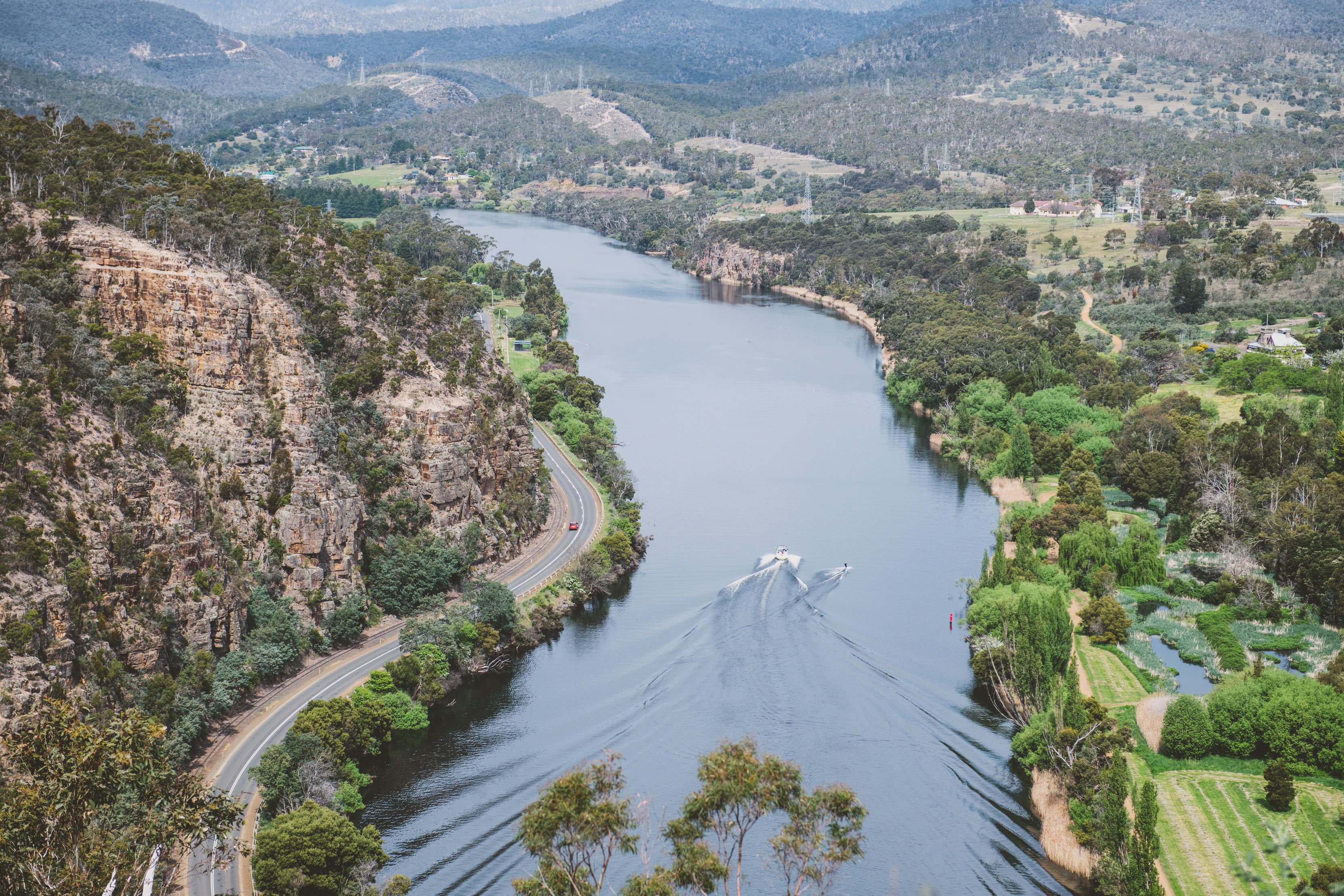 An aerial view of a wide river, with two boats travelling down it. The landscape is hilly and bushy, a road follows the river on one bank and some landscaped gardens are on the other.