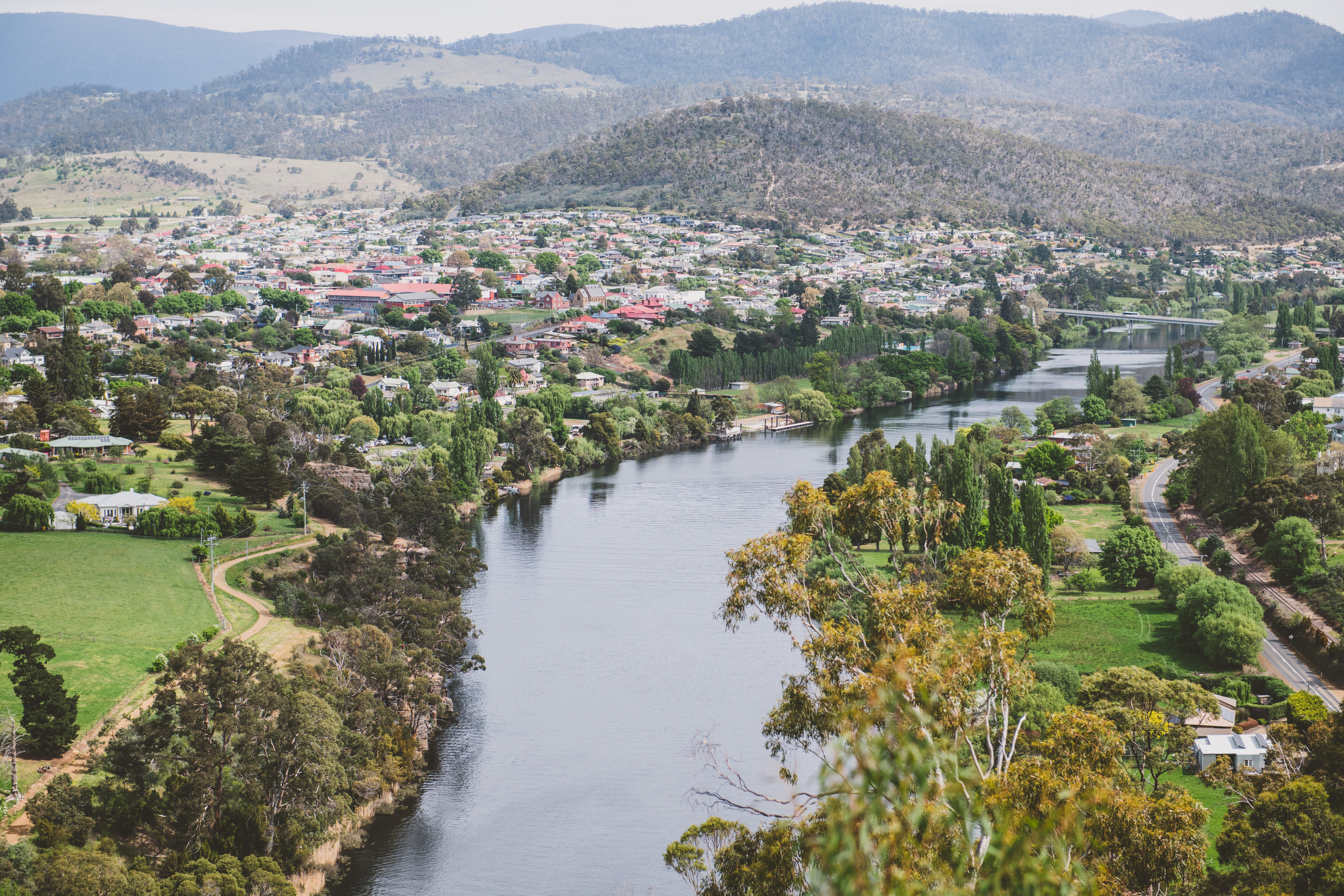 An aerial view of a river running through a small town. The landscape is dotted with trees, bush and houses, and mountains rise up behind.