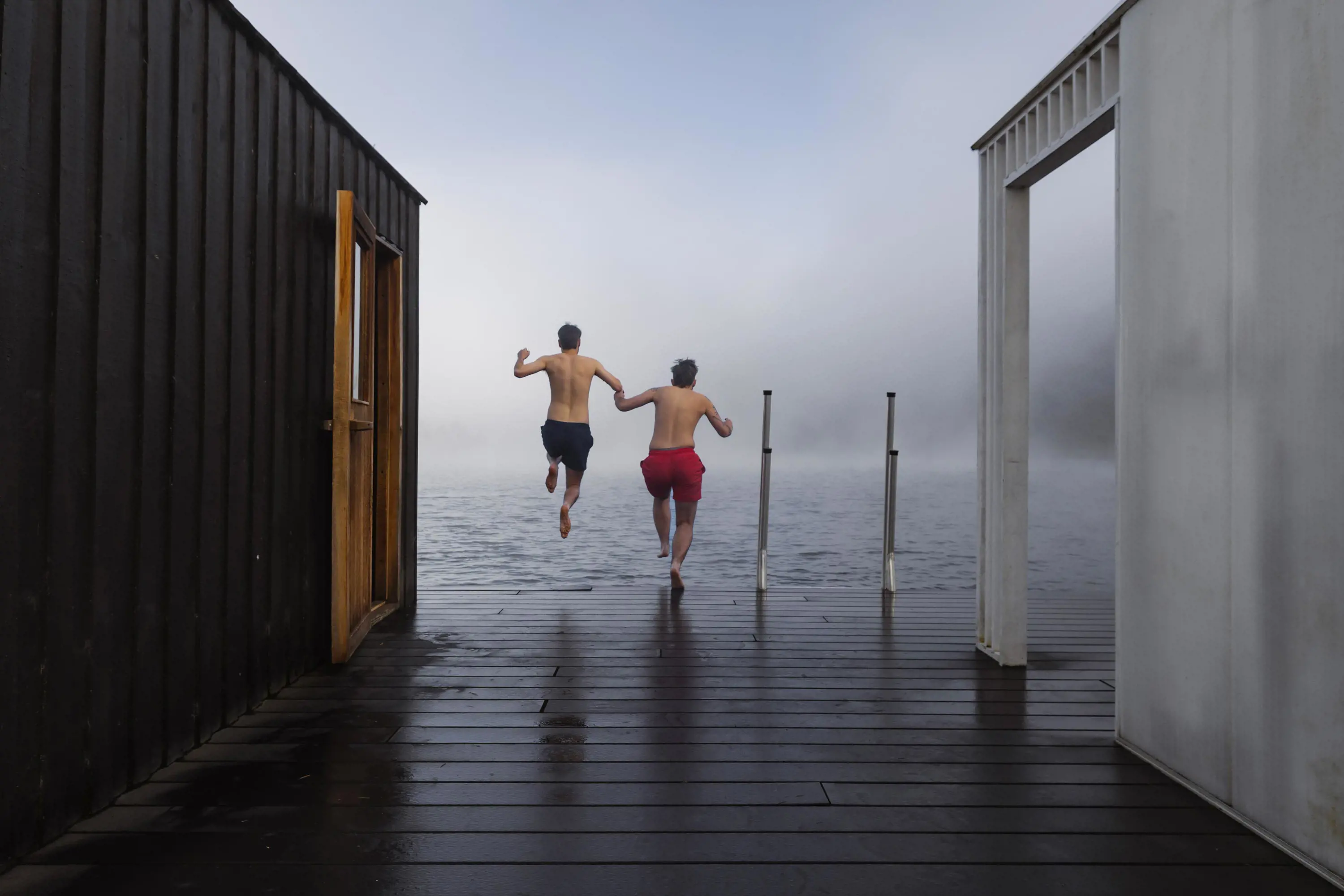 Two young men holding hands jump into the cold waters of a lake from the decking in front of a sauna.