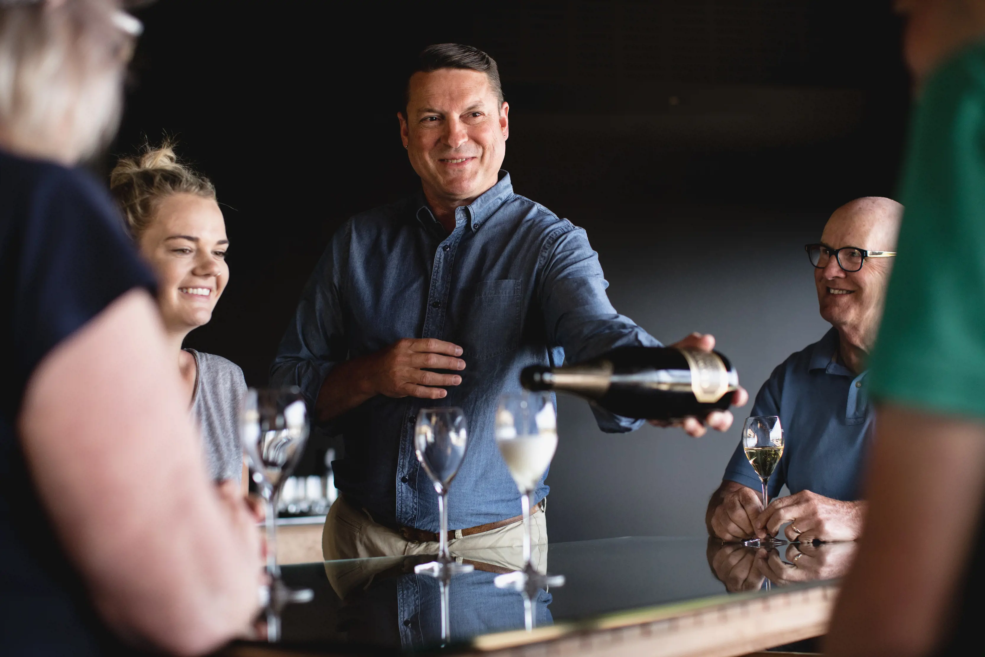 A group of people around a bar, holding wine glasses. One man pours wine into a glass from the bottle.