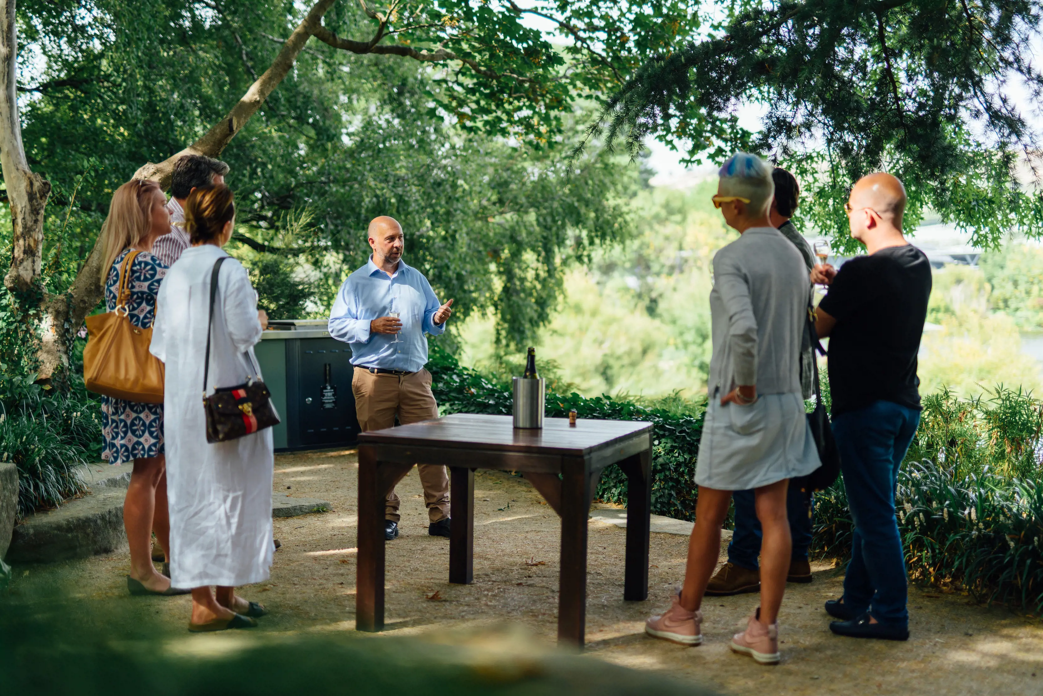 A group of people holding wine glasses stand outdoors around a table with a wine bottle on it, listening to a man speak.