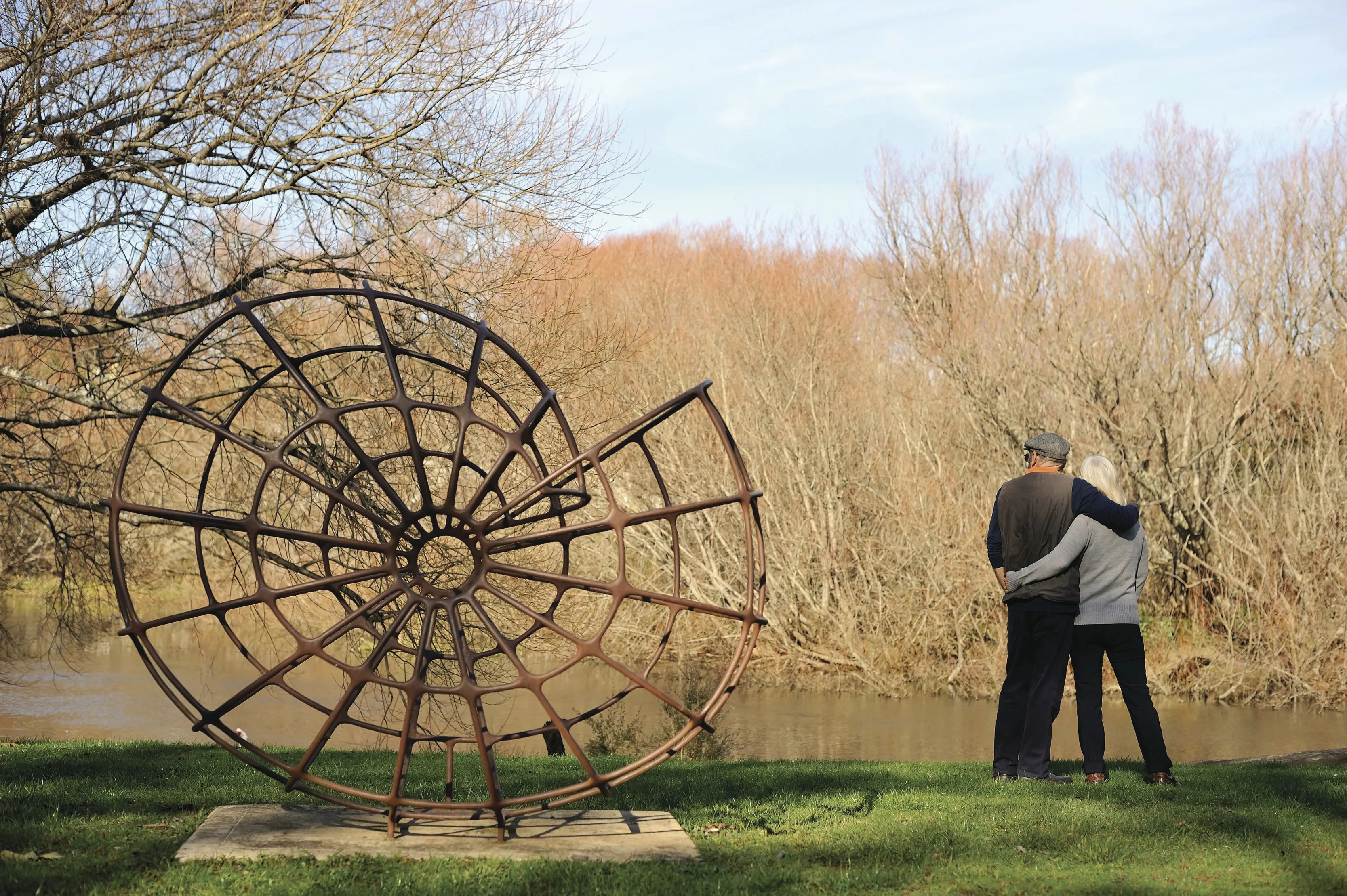 A rusted metal sculpture of a spiral shape sits on the grassy bank of a river. Two people have an arm around each other standing nearby.