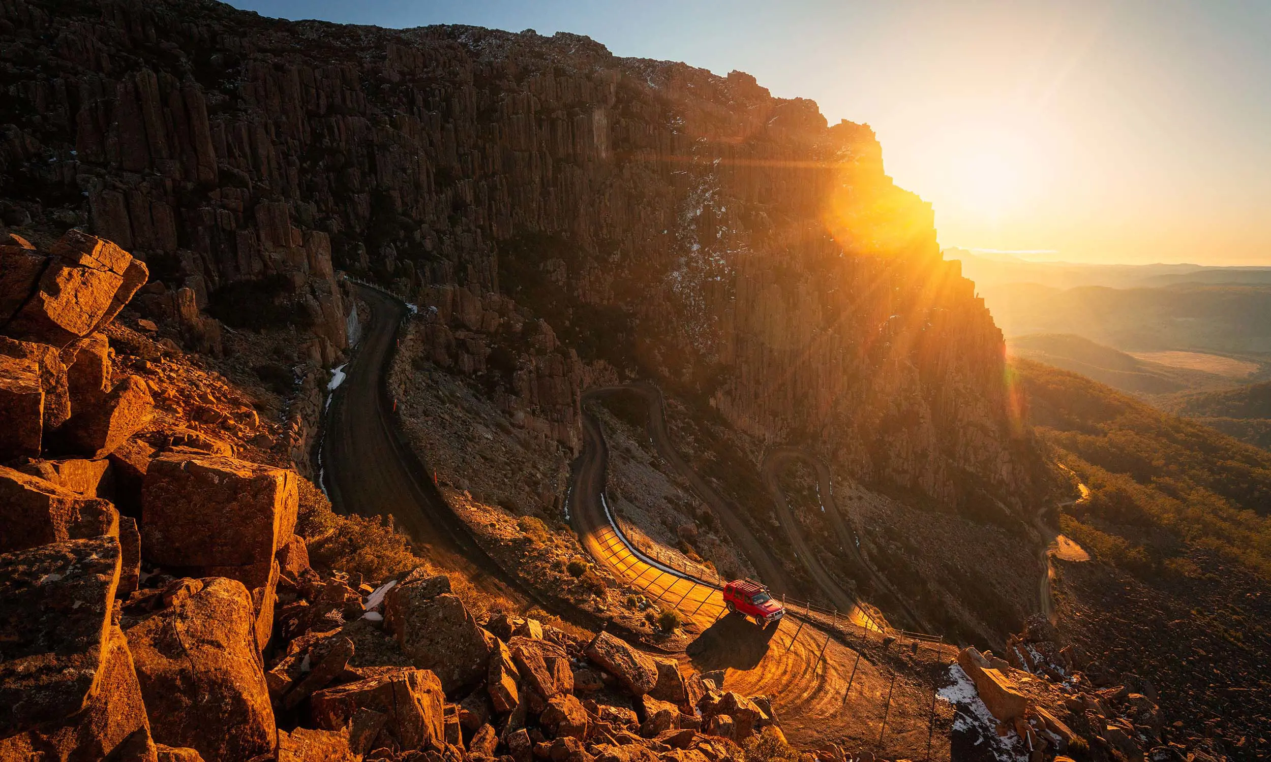 A dramatic rocky cliff illuminated by the setting sun, with a winding dirt road and a parked vehicle at its base. The warm sunlight highlights the rugged terrain and creates long shadows, emphasizing the texture of the rocks.