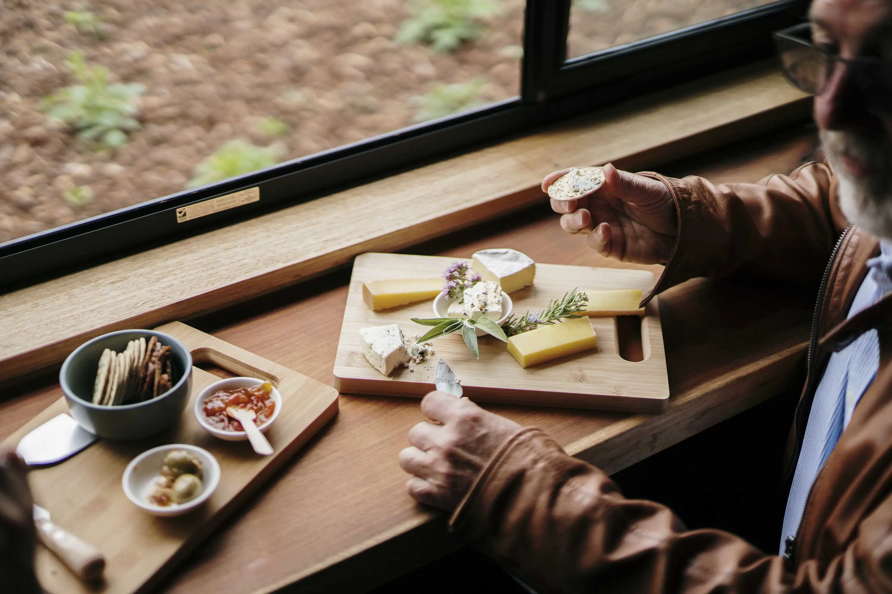 A man sits at a bench eating a cheese platter with jams, olives and crackers.  