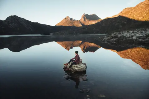 Person sitting on a rock in the middle of a lake amongst Cradle Mountain. The jagged contours of Cradle Mountain surround the image.