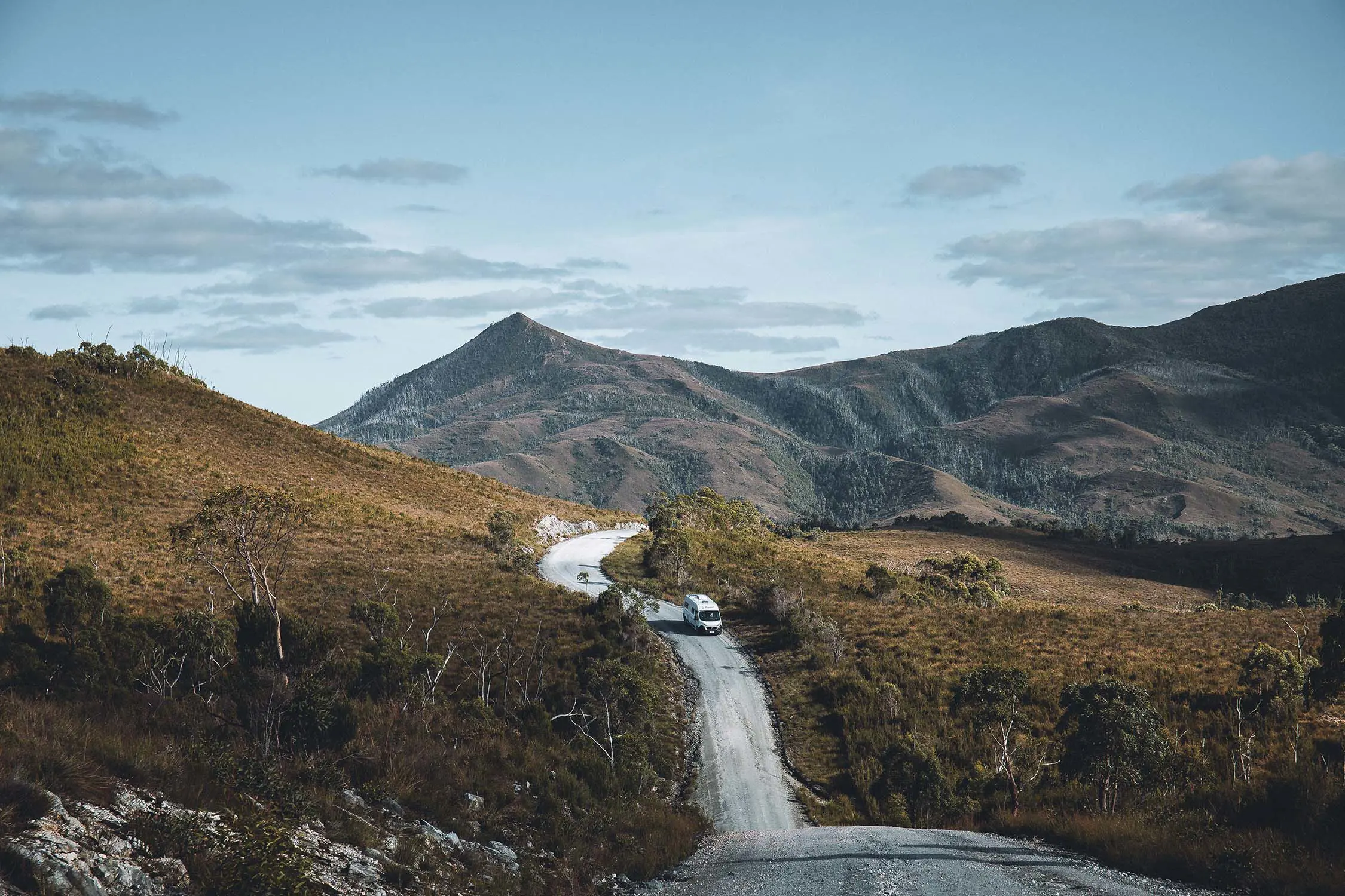 A van drives down an unsealed gravel road, which winds up and down hills covered in dense alpine bush.