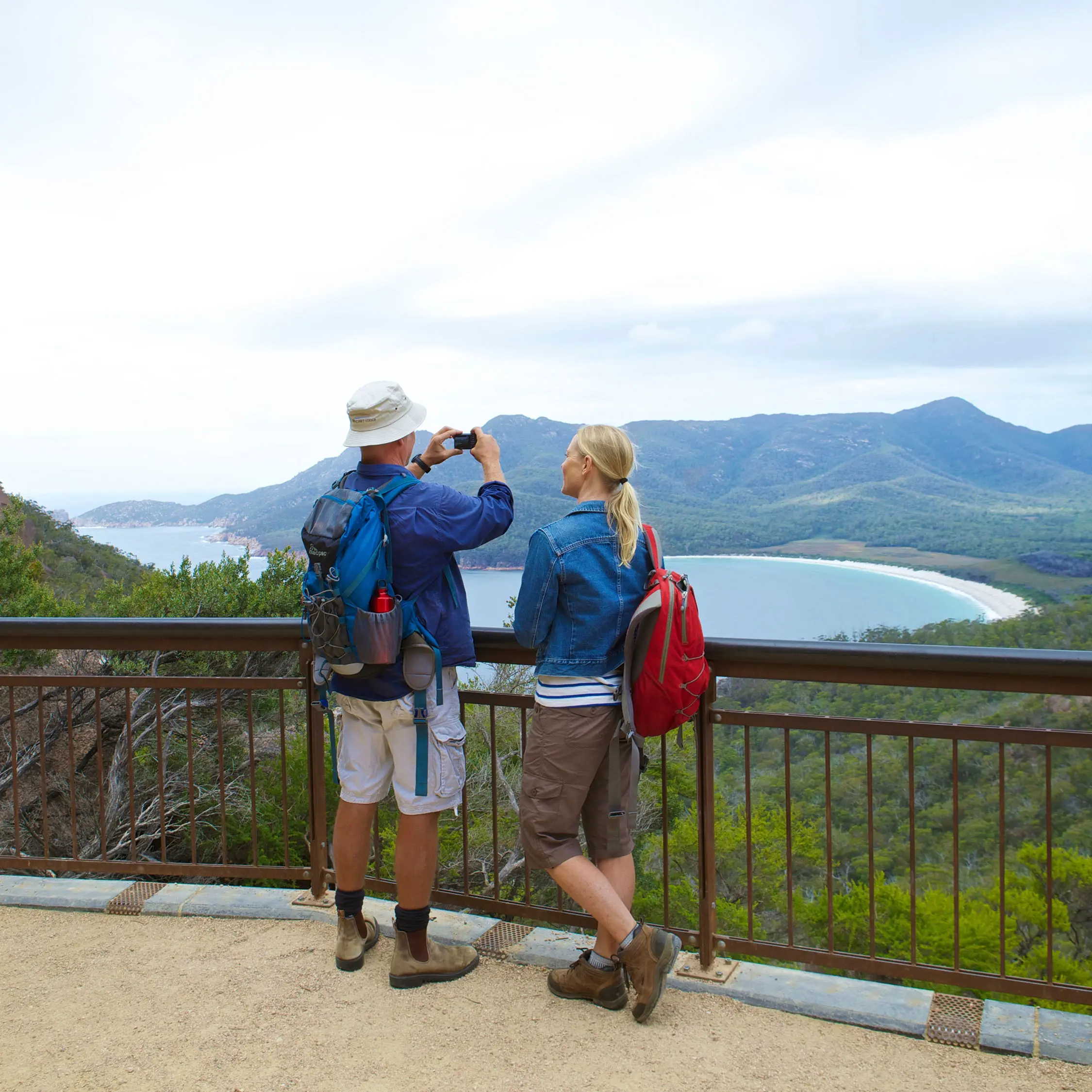 Two people in hiking clothes and day packs stand looking out over Wineglass Bay from a lookout platform, taking a photo.
