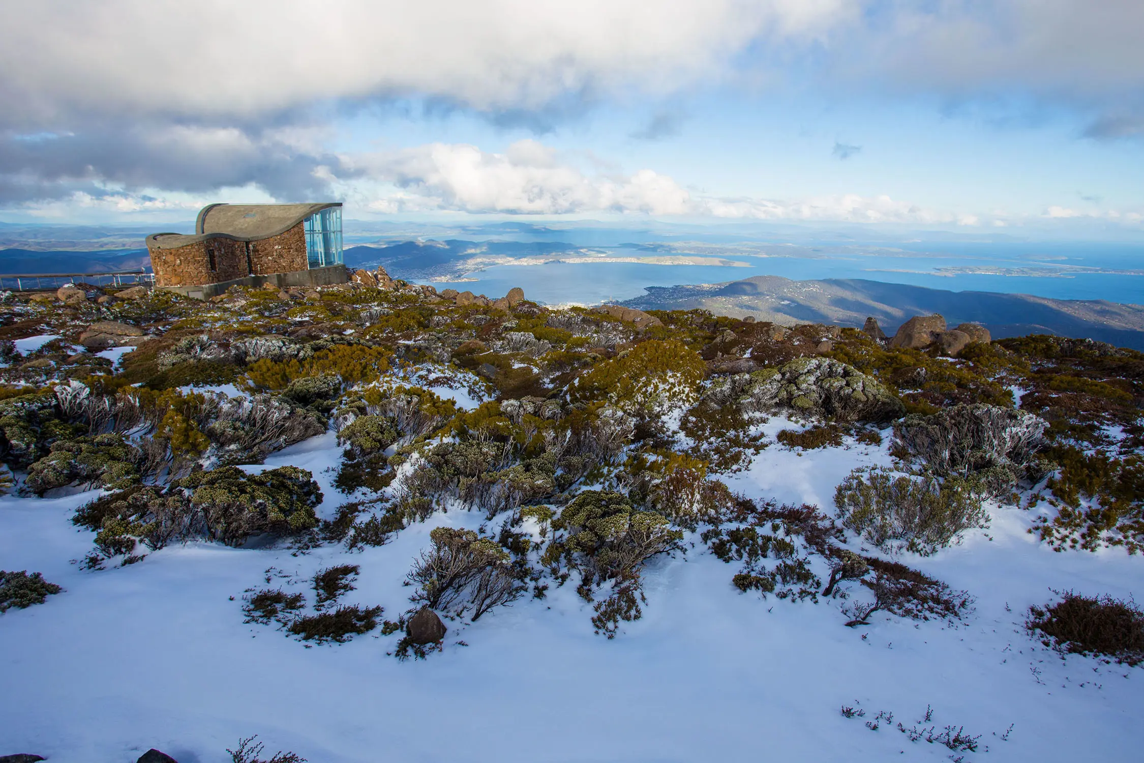 A panoramic view from a mountaintop showing a modern building with a curved roof, rocky terrain with patches of snow, vegetation, and distant valleys under a cloudy sky.