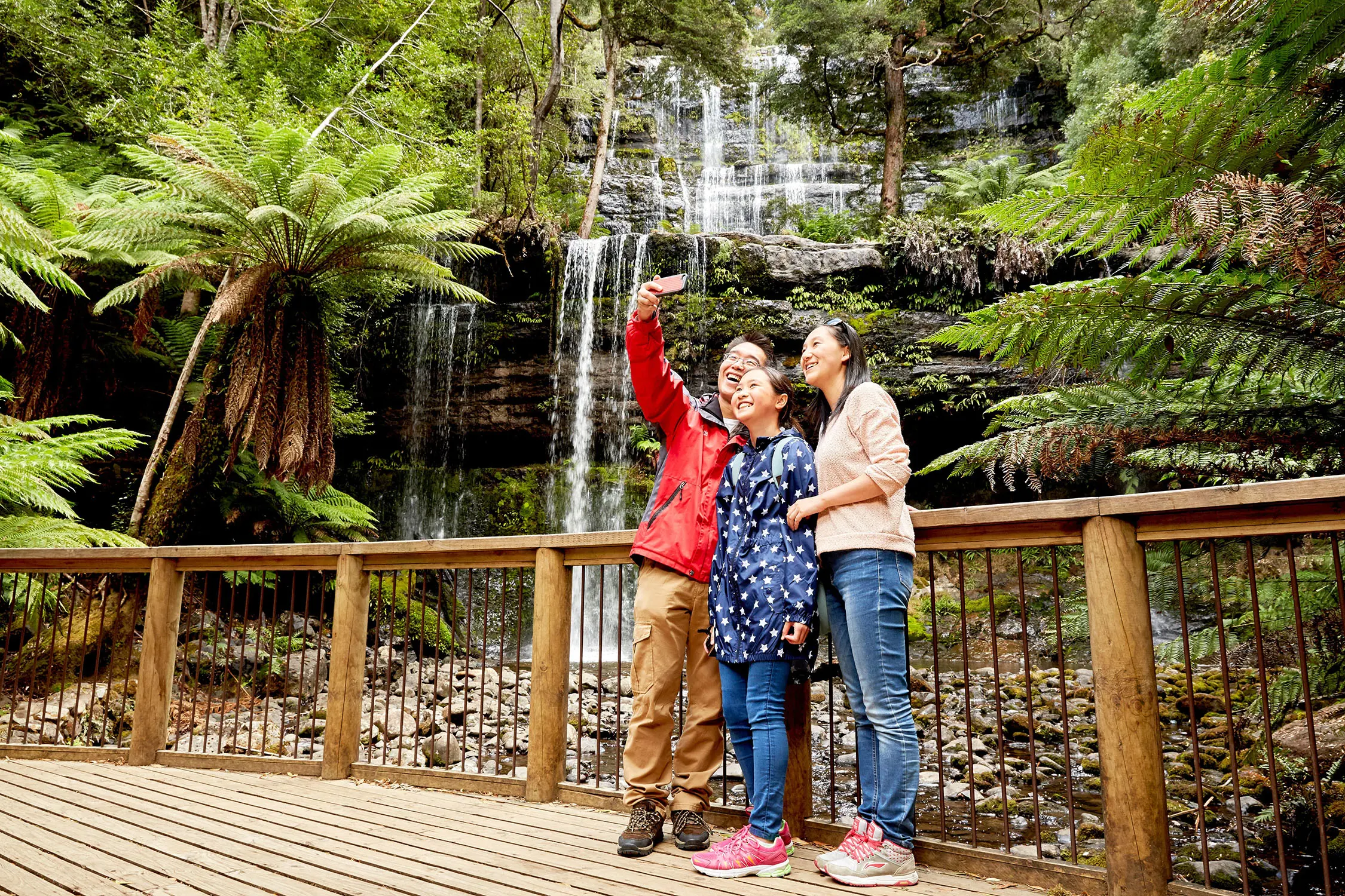 A family stands on a wooden boardwalk in front of a lush rainforest waterfall, with one person holding out a phone to take a selfie of them.