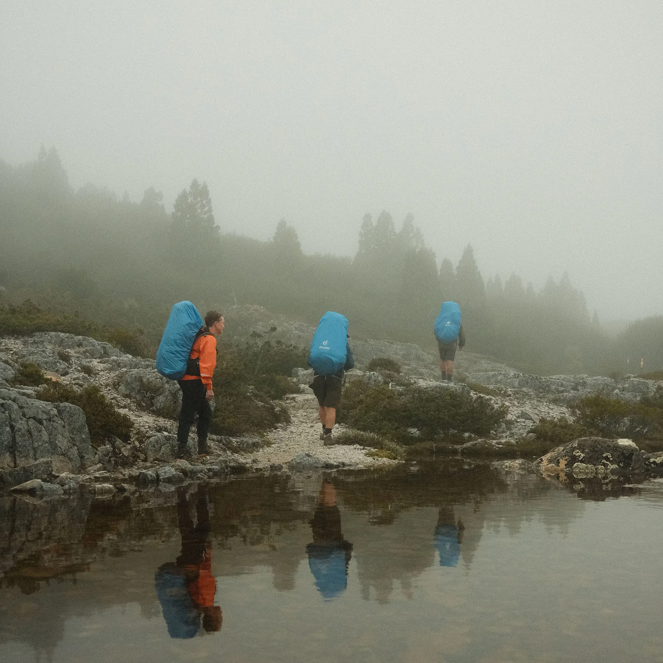 Three people in hiking gear and large covered backpacks hike through a foggy landscape next to a still lake.