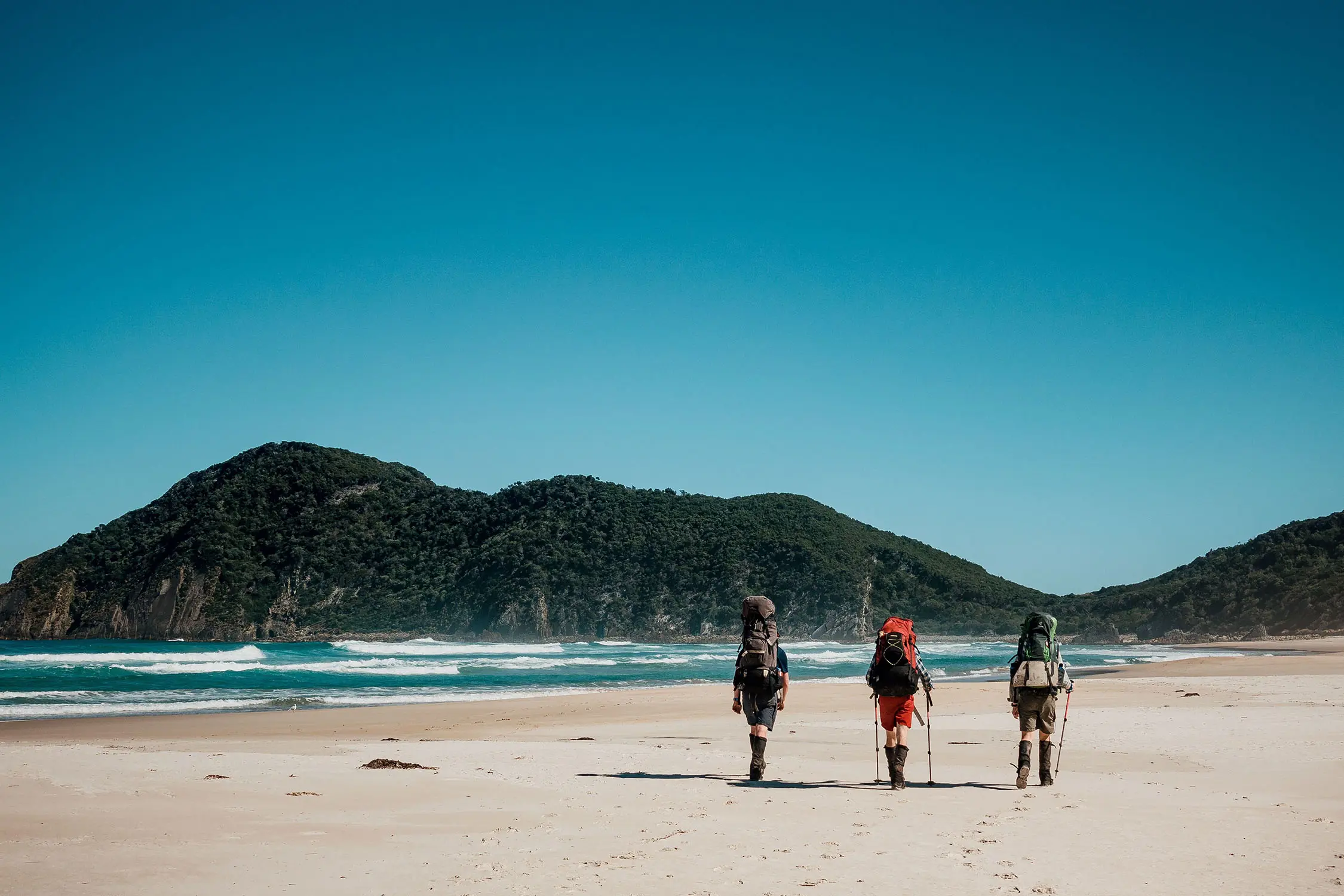 Three people wearing hiking gear, gaiters and packs, and carrying walking poles, walk across a wide open sandy beach.