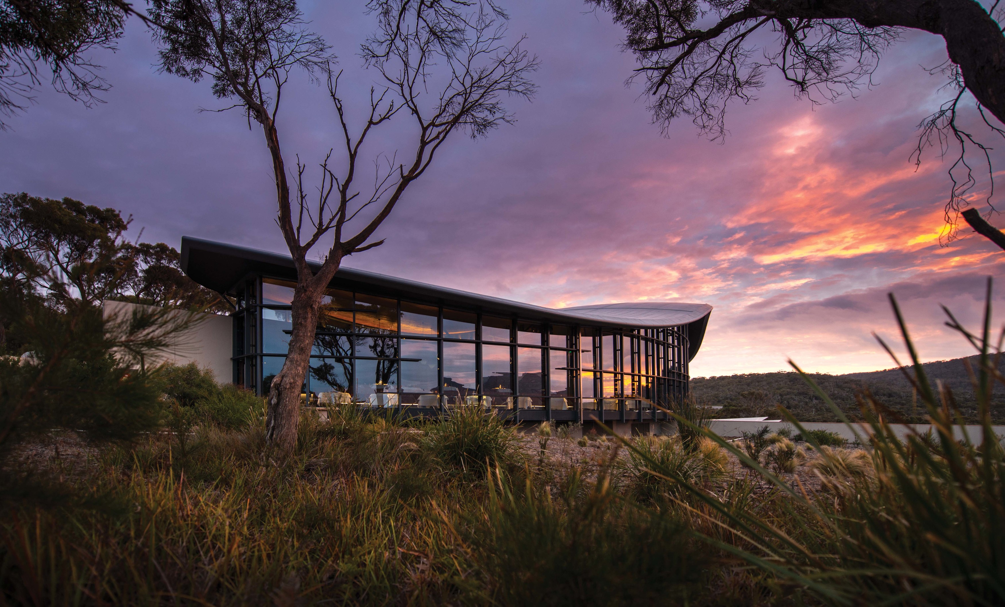 Image of glass house surrounded by grass. Mountain view in the background. Pink and purple sunset.