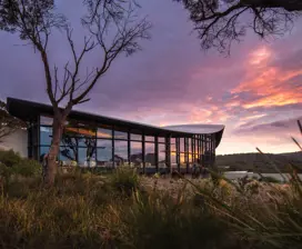 Image of glass house surrounded by grass. Mountain view in the background. Pink and purple sunset.