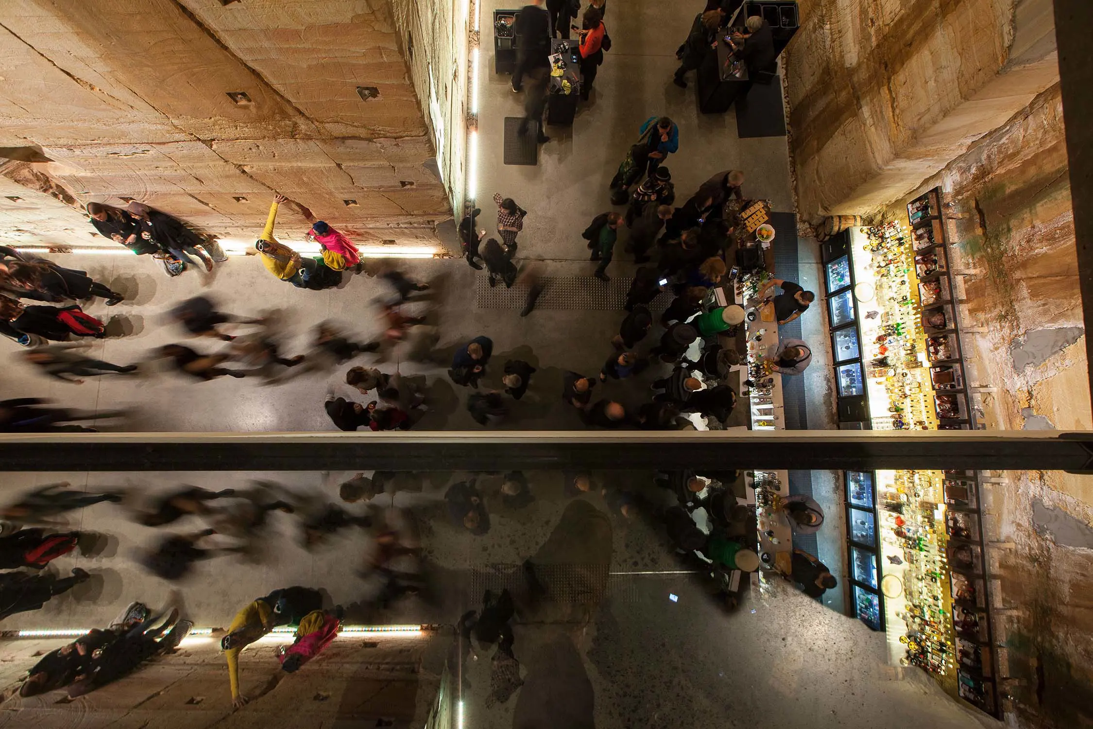 A top-down view of a busy underground T-shaped walkway, with cut-stone walls and streams of people walking through, their motion slightly blurred. A long bar is at one end with bartenders serving a queue of patrons.