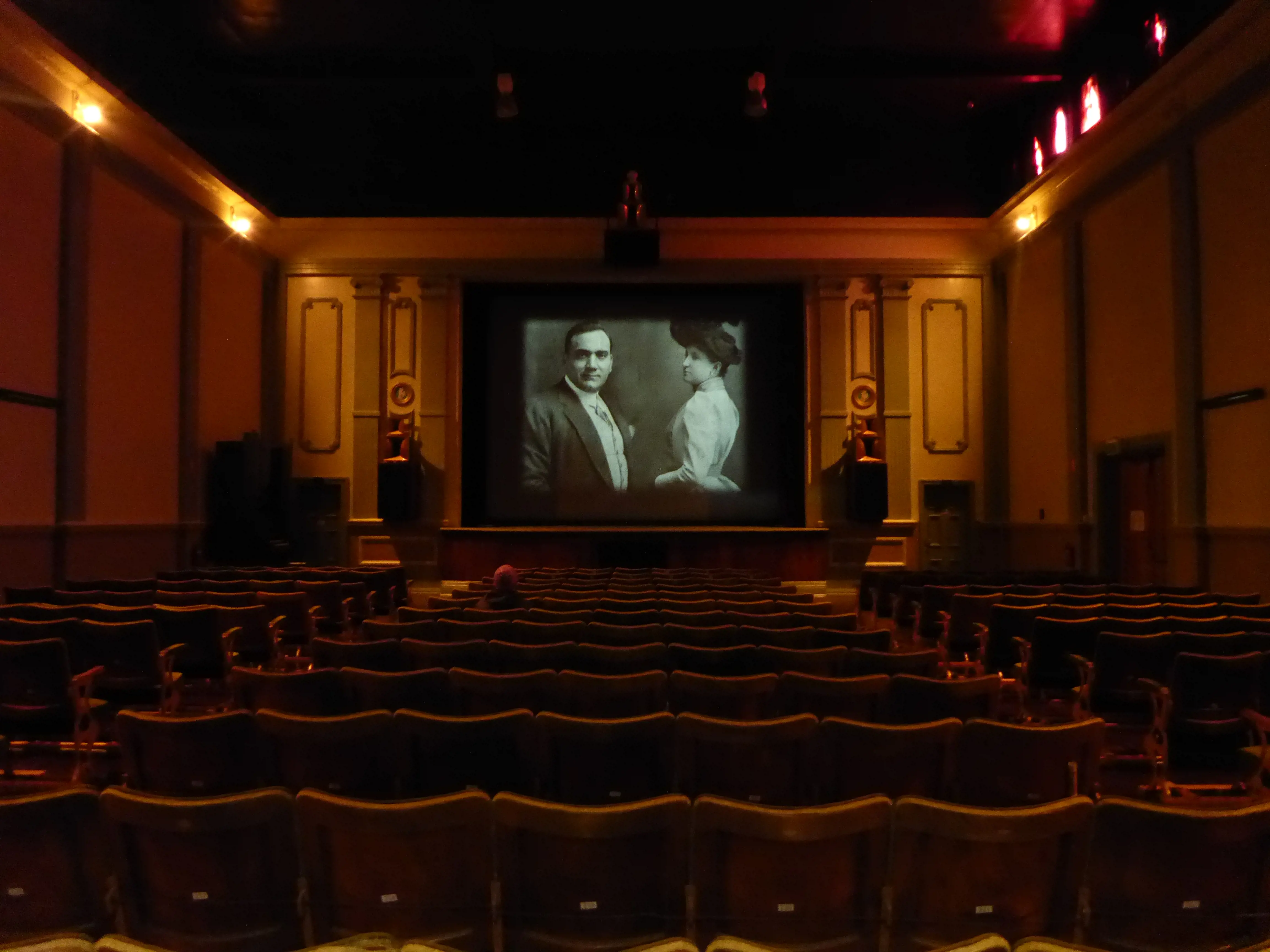 A black and white film plays in a warmly lit art-deco style theatre with high back, red velvet seating in rows.