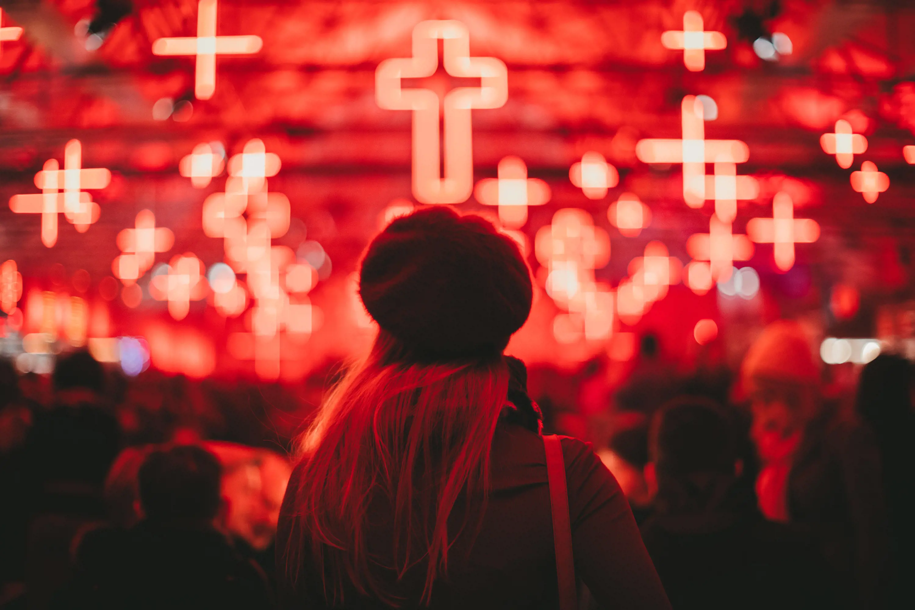 The head and shoulders of a woman facing away from the camera. The background is out of focus, with a red hue and many hanging neon crosses.