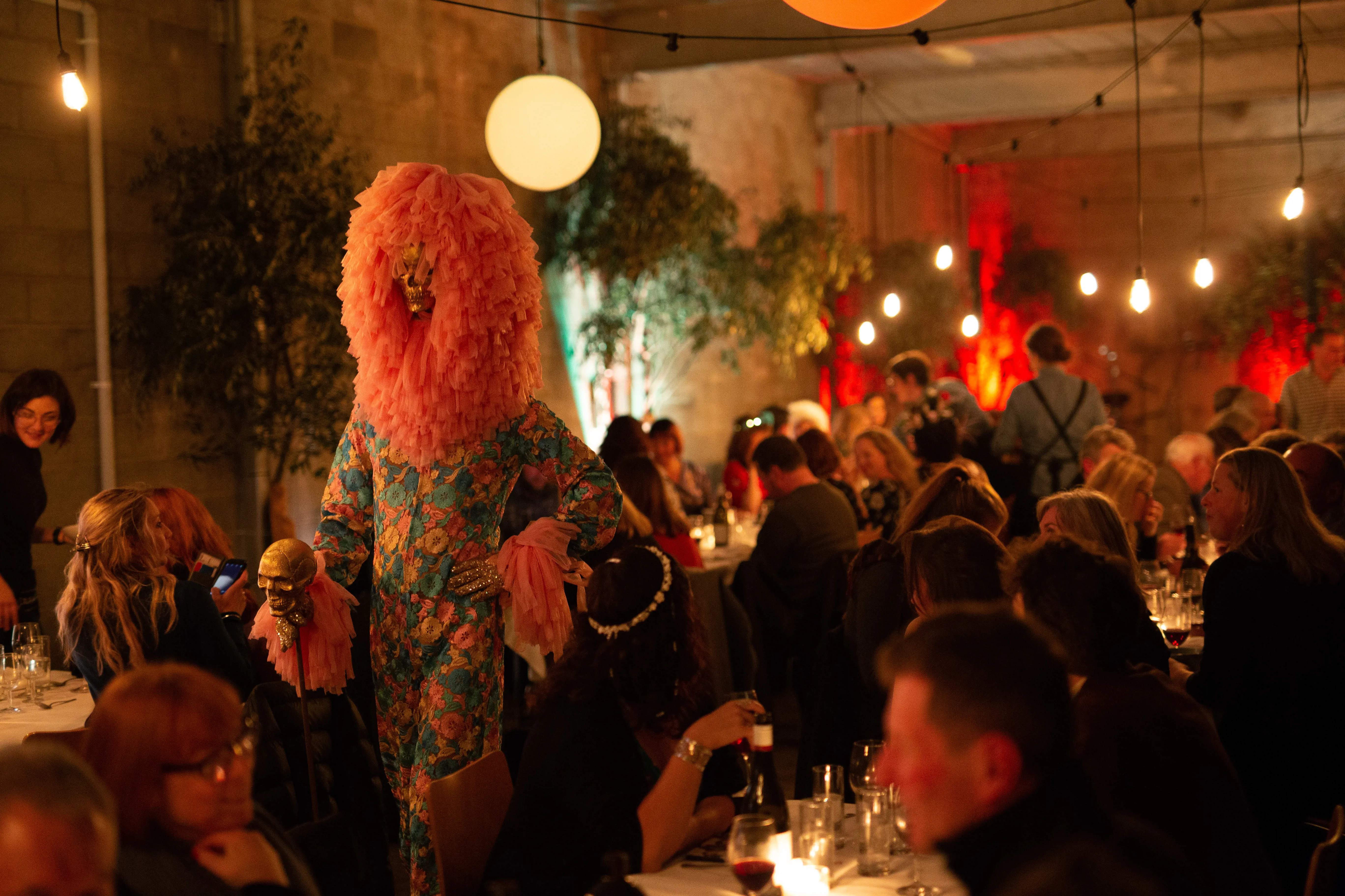 A strangely costumed person walks among busy dinner tables at an event.