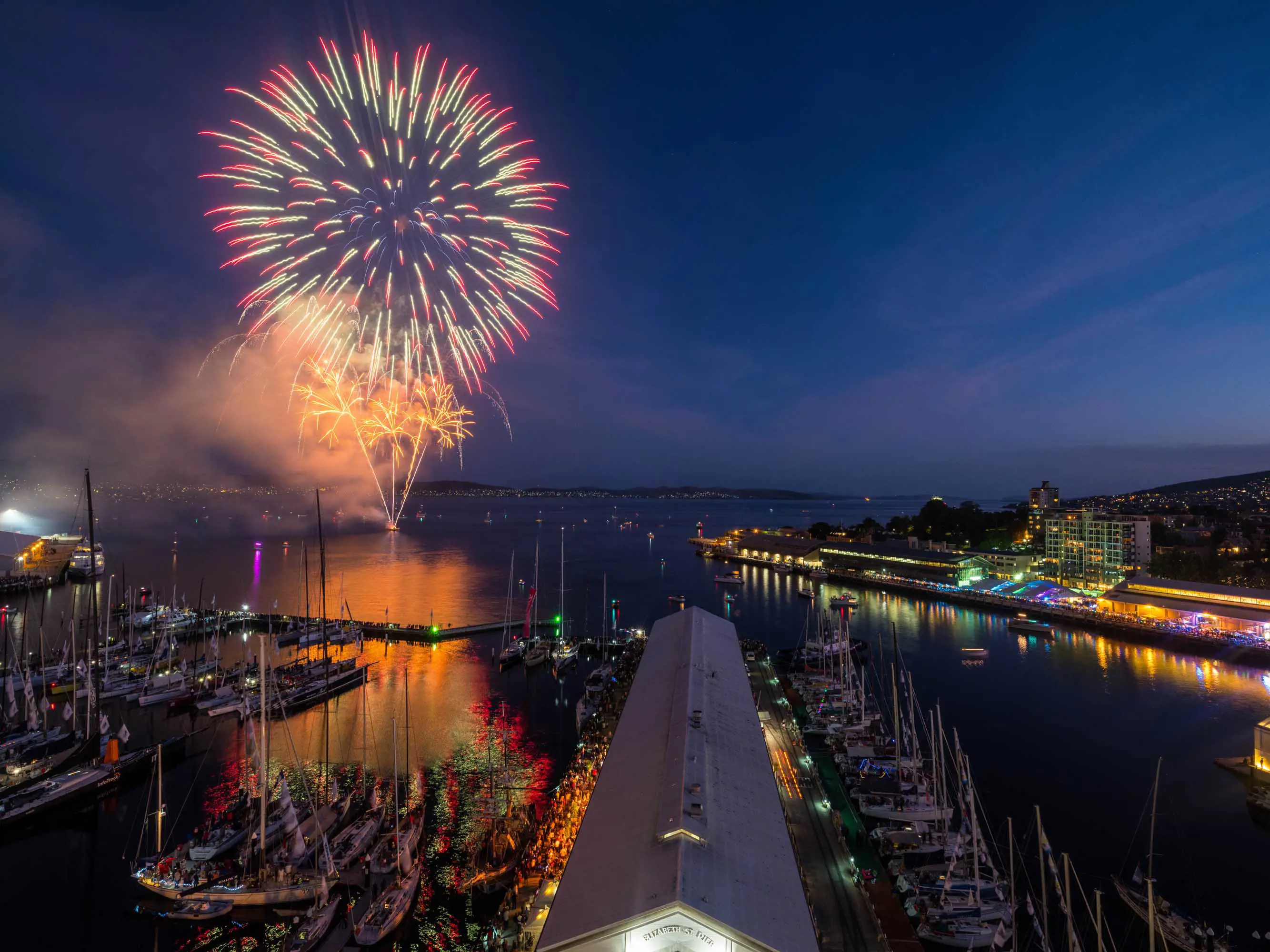 A large fireworks display at the waterfront of Hobart is reflected in the calm waters of a river.