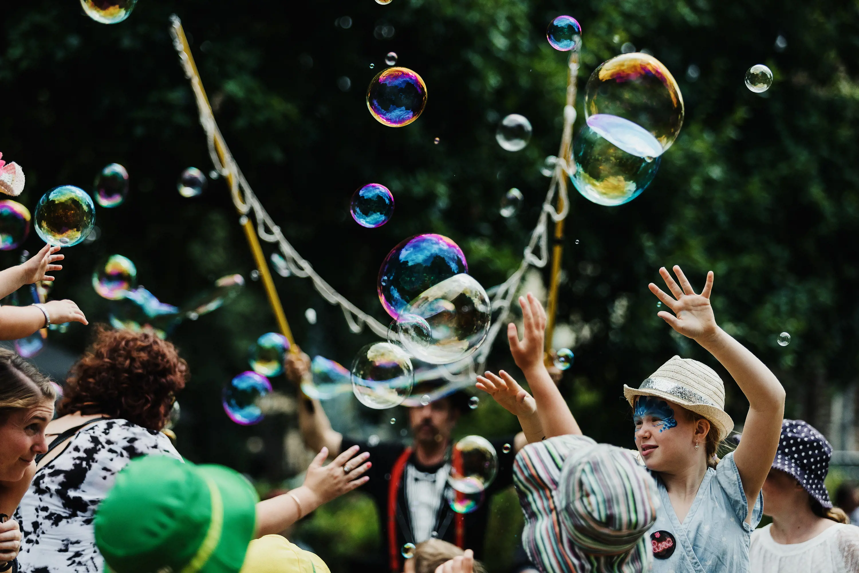 A group of adults and children reach towards floating bubbles, which are created by a man holding up two long sticks with strings of loops hanging between them.