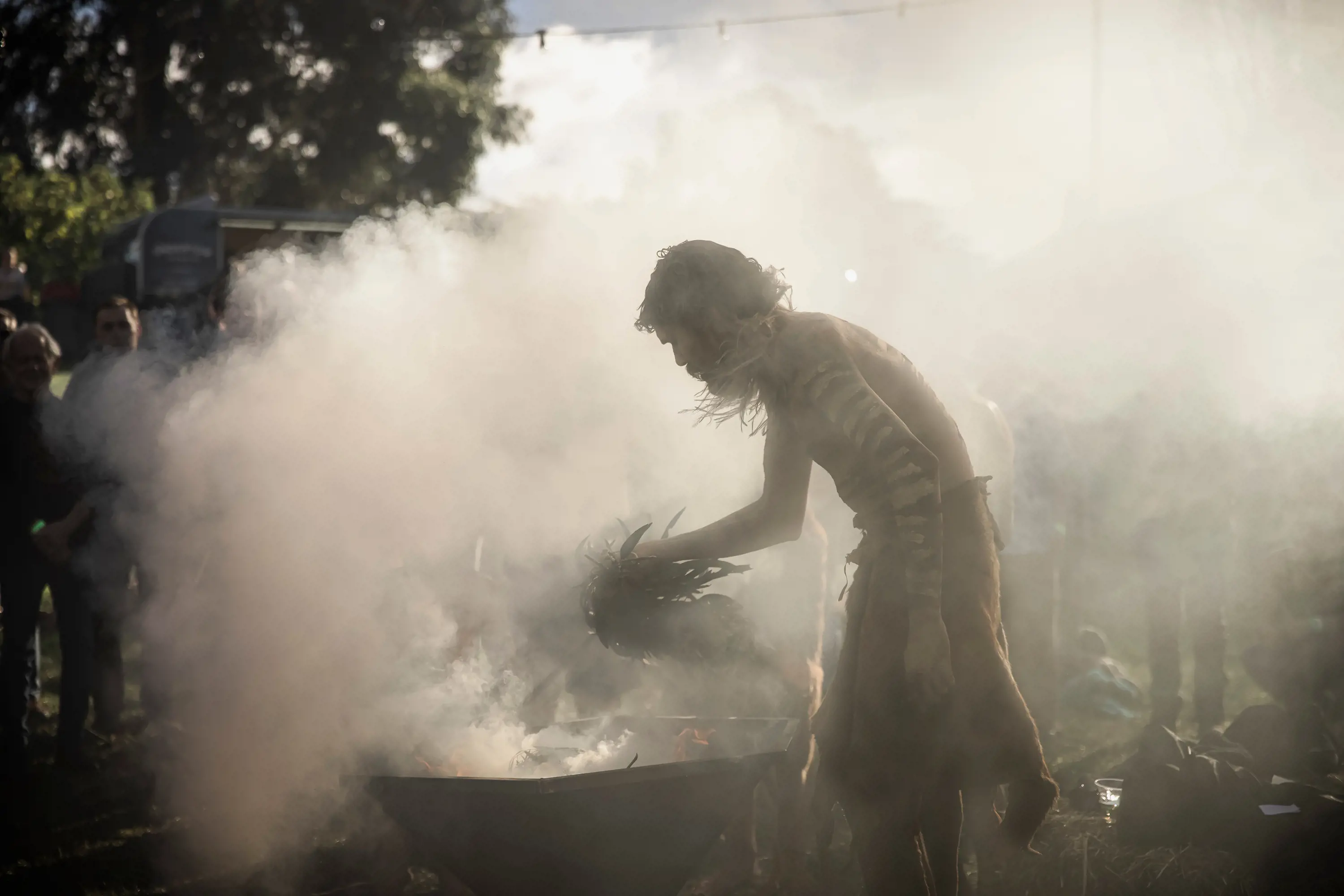 A man with indigenous traditional marks painted down his arms and face holds a handful of leaves over a very smoky fire bowl.