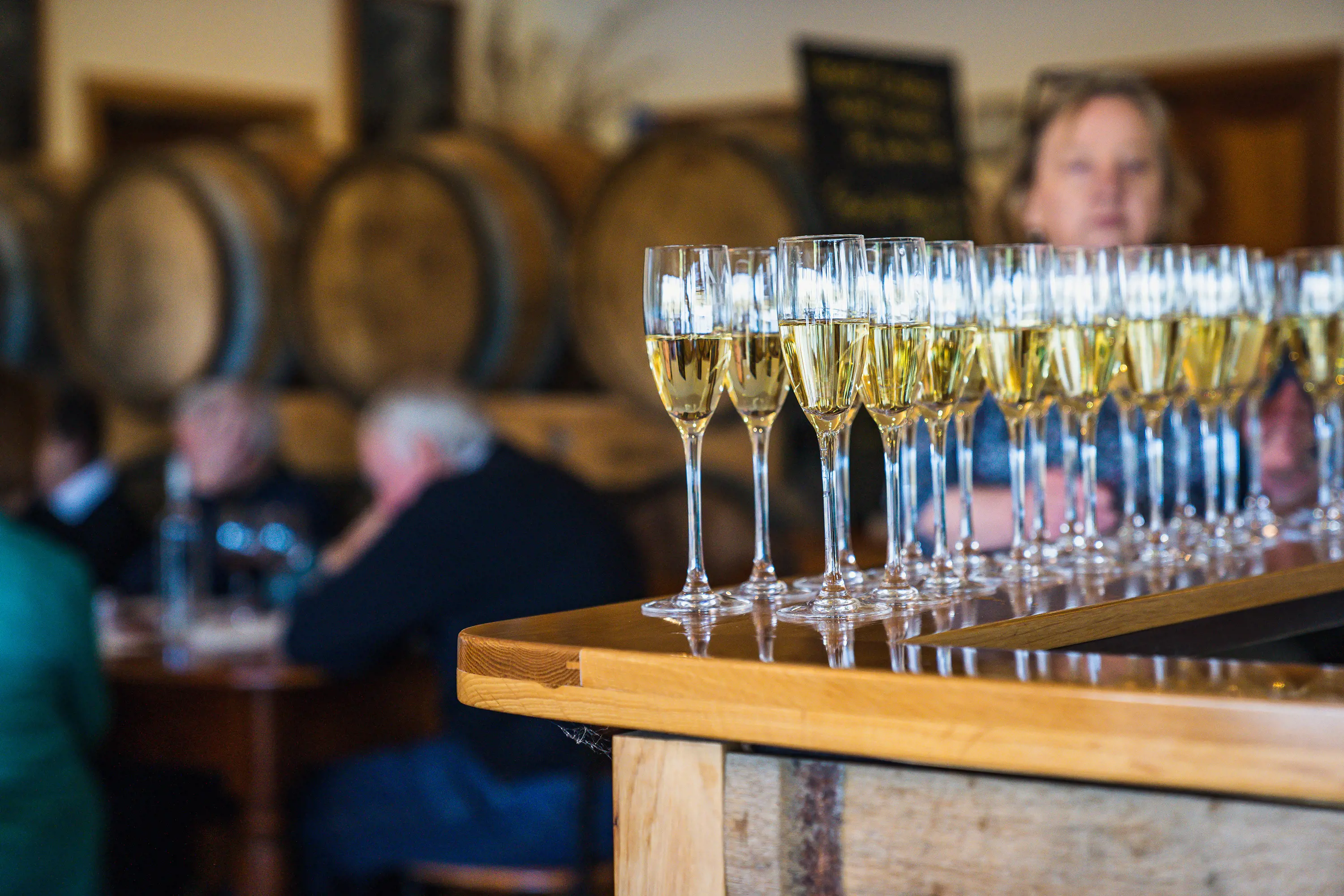 Lines of half-filled champagne flutes stand on the corner of a wooden bar. In the background are huge barrels and people sitting at tables dining.