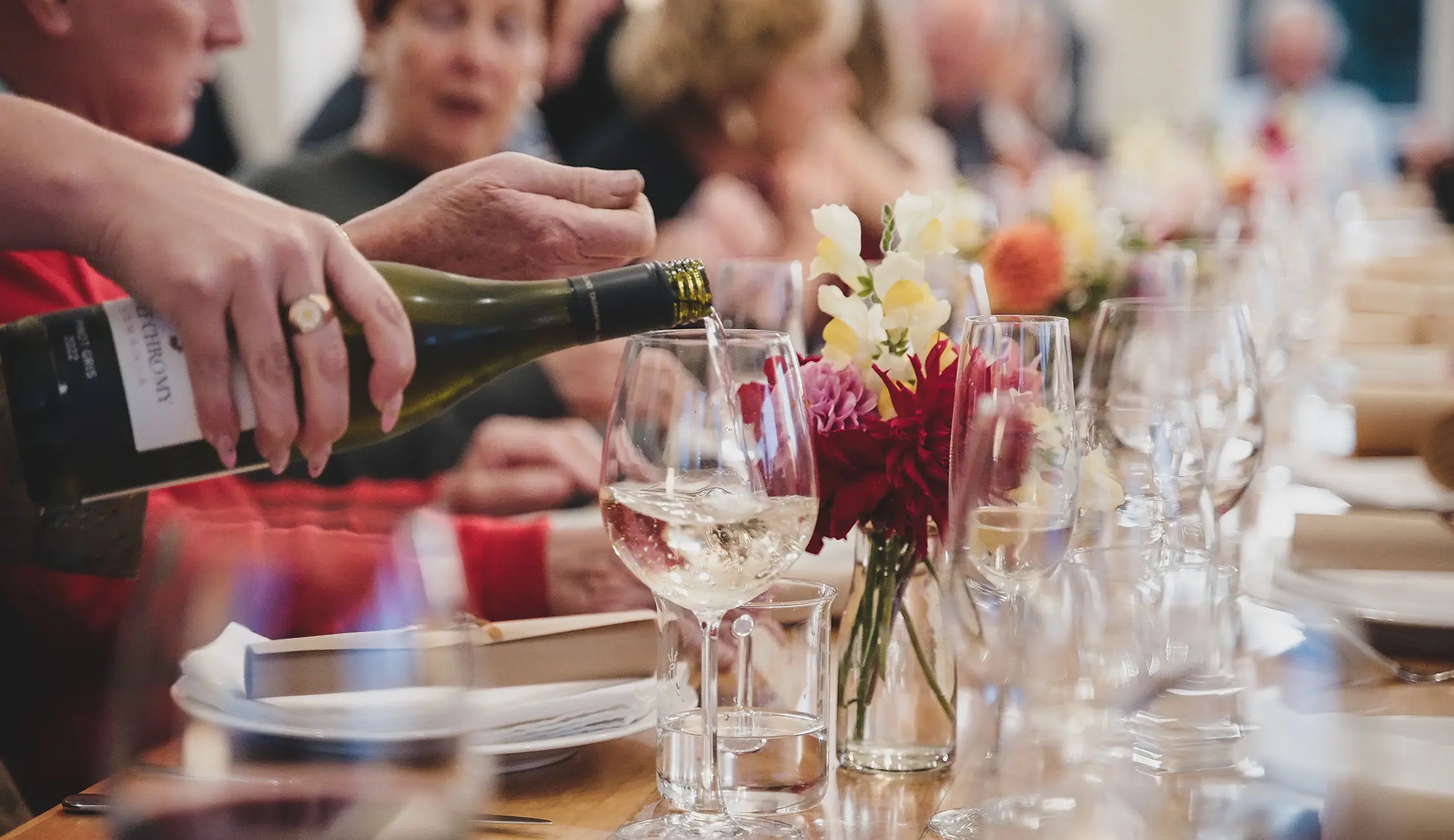 A close-up of a long table lunch. The table is set with flowers, plates and glasses, and a hand is visible pouring a bottle of white wine into a glass.