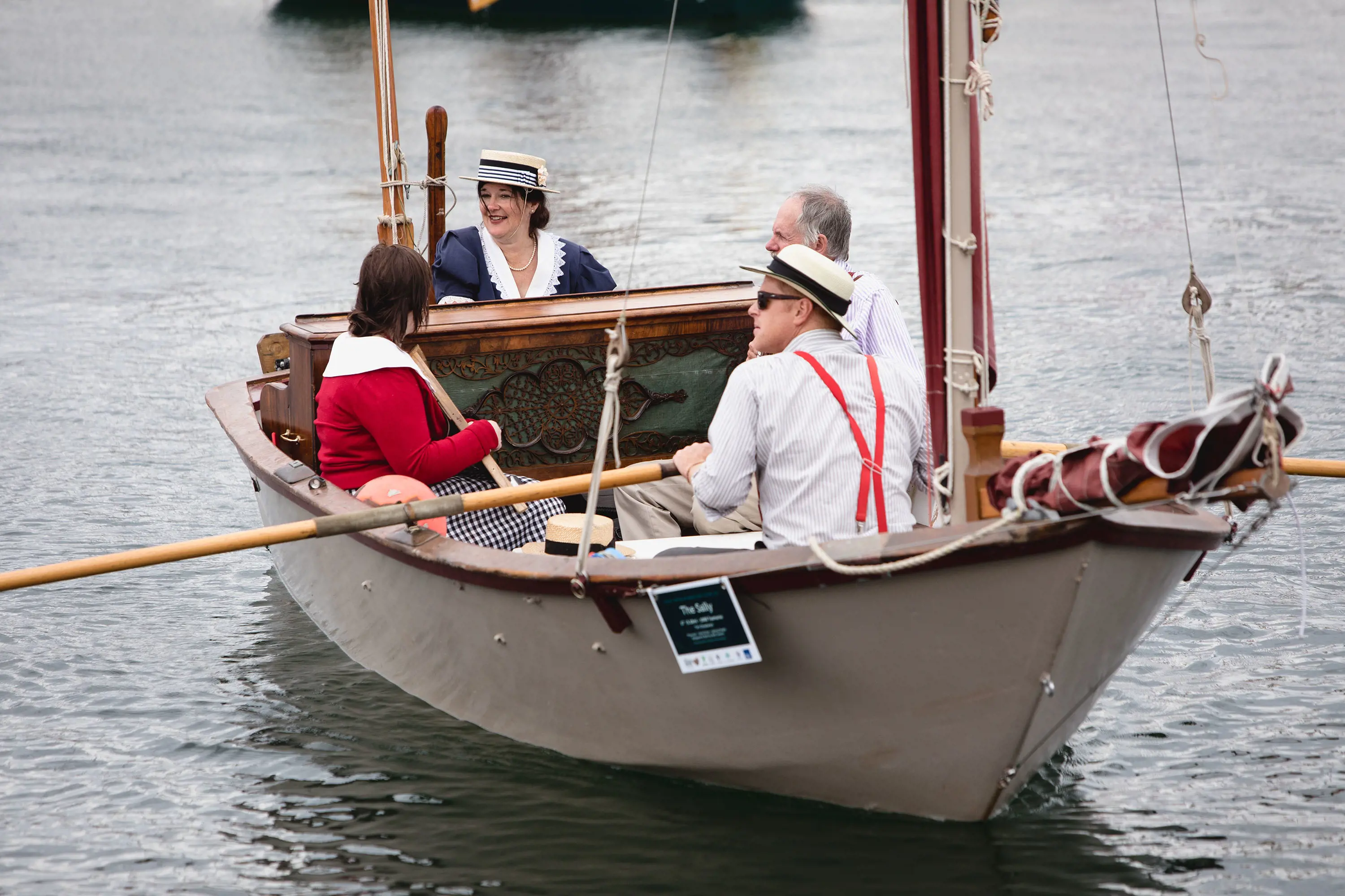 Four people in vintage boating outfits and hats sit onboard a traditional wooden dinghy boat, with oars and a small mast.