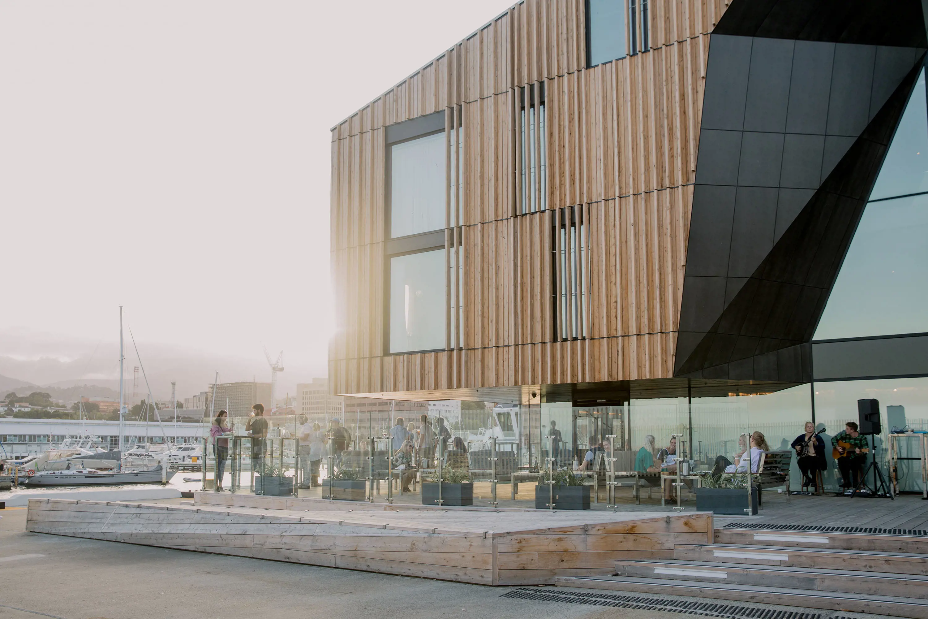 A modern wood-panelled hotel building next to a harbour. On the deck behind glass panels, people sit at tables.