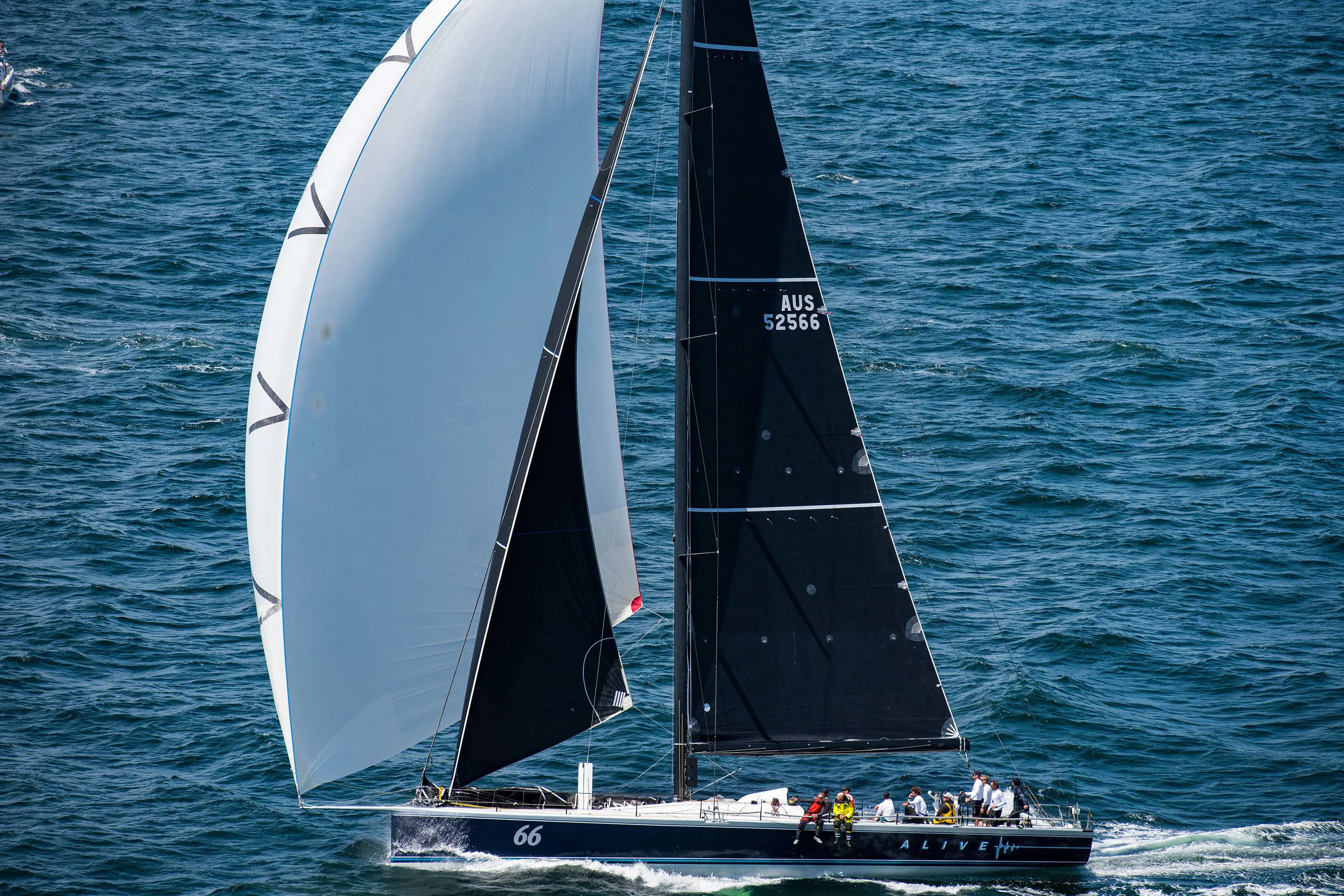 A sailing yacht travelling on blue-grey water. People onboard look small compared to the large, billowing black and white sails.