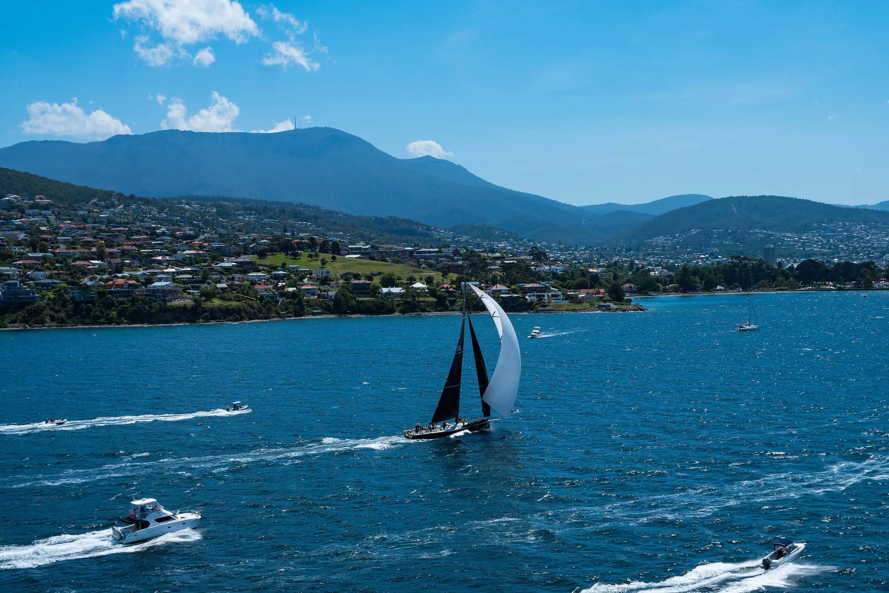 A sailing yacht and a few other small boats travelling up a wide, blue river. On the bank is a built up suburb, with a mountain range rising up into the blue sky.