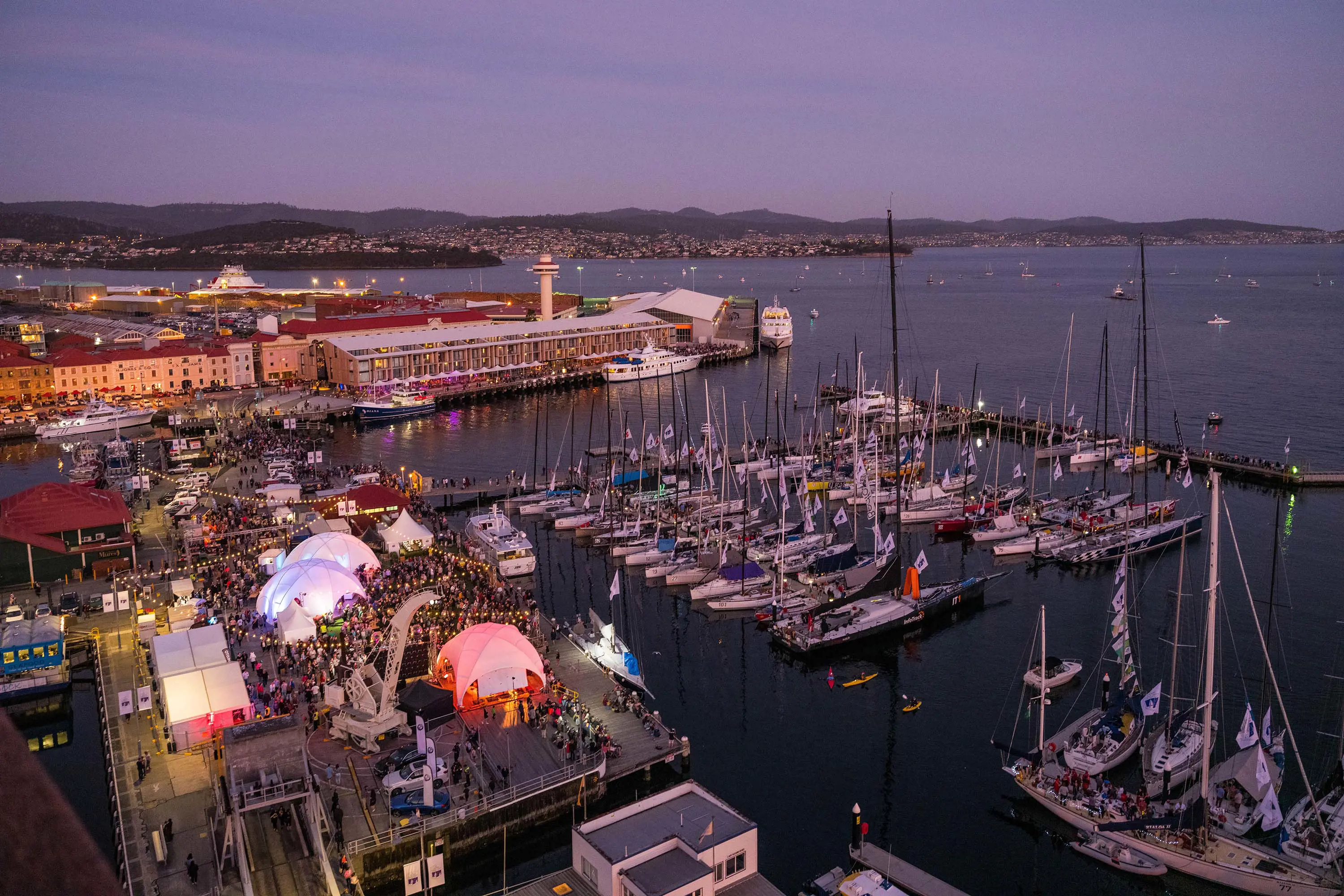 An aerial view of a busy harbour. Many boats are moored in the marina, and on the wharf there are marquees, lights and crowds of people.