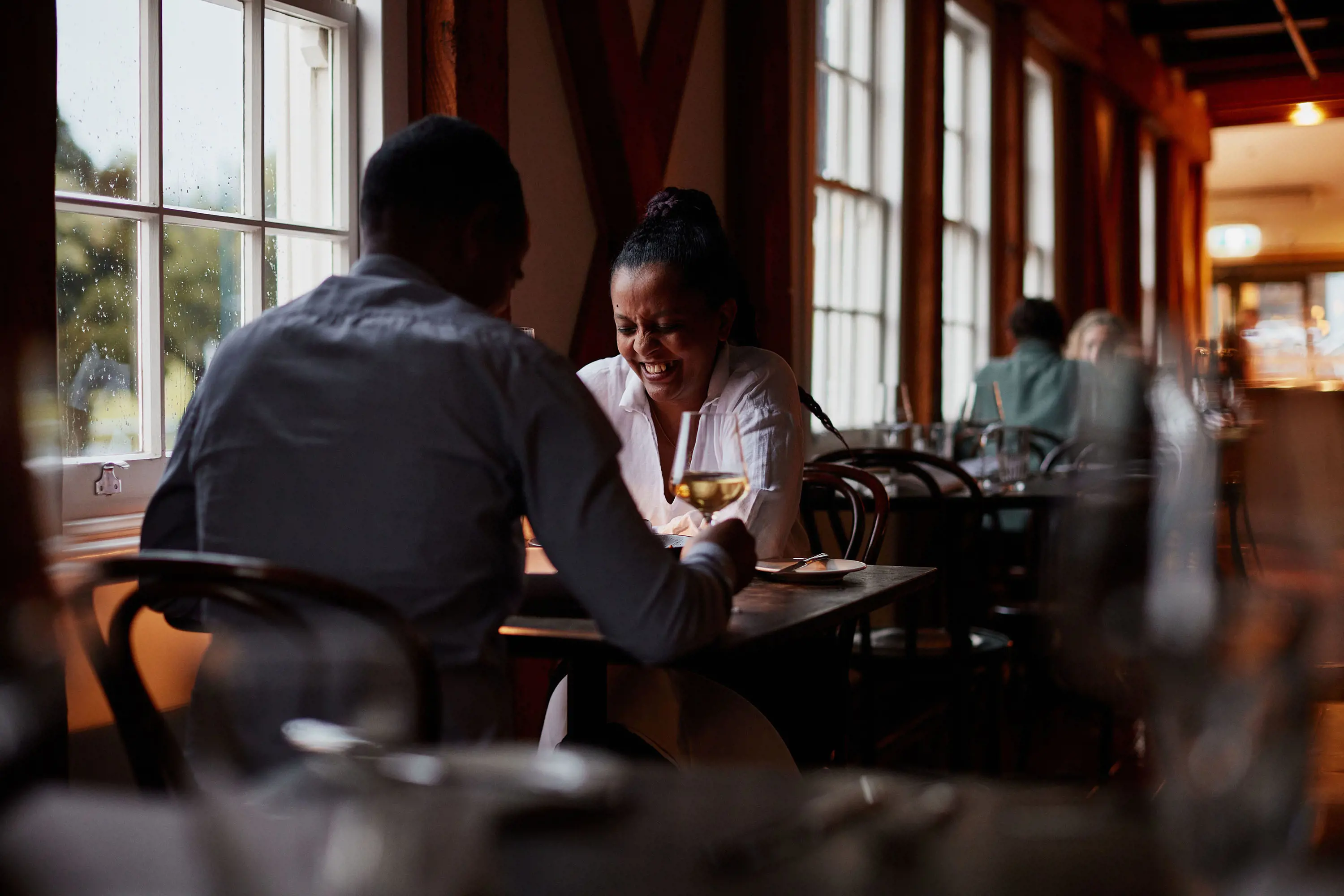 Two people sit at a small intimate dining table in a restaurant, drinking cocktails and laughing. The lighting is low and moody, and the window panes are dotted with raindrops.