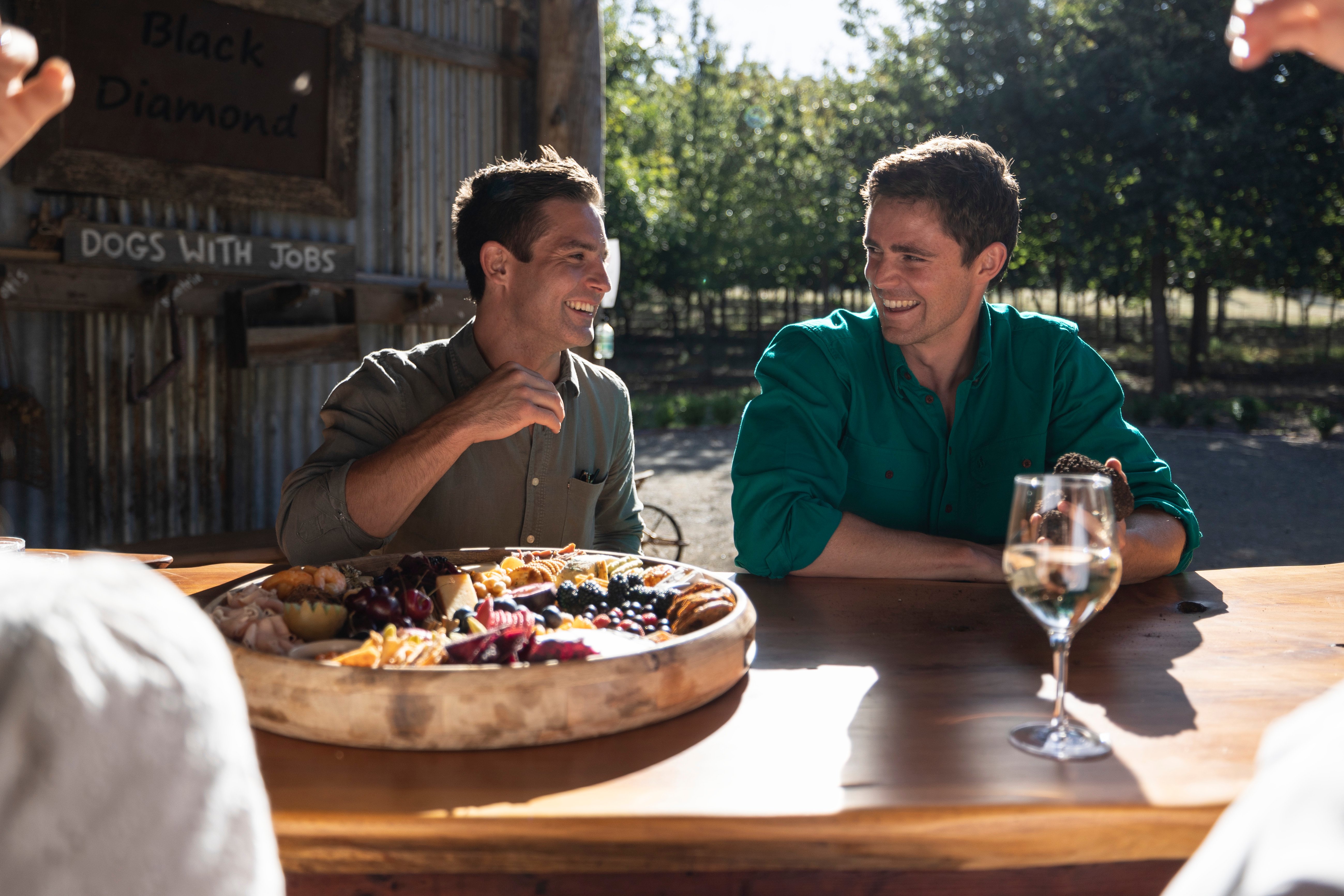 Two friends enjoy a platter of cheese and fruit at a long wooden table.