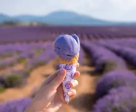 A person's hand, holding up a lavender icecream cone. The out-of-focus background is a long field of purple-hued small shrubs.