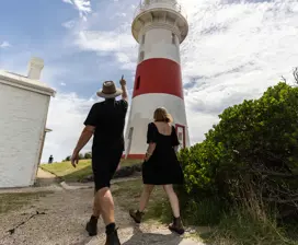 A guide and a tourist walk along a gravel path below a tall, red and white lighthouse.